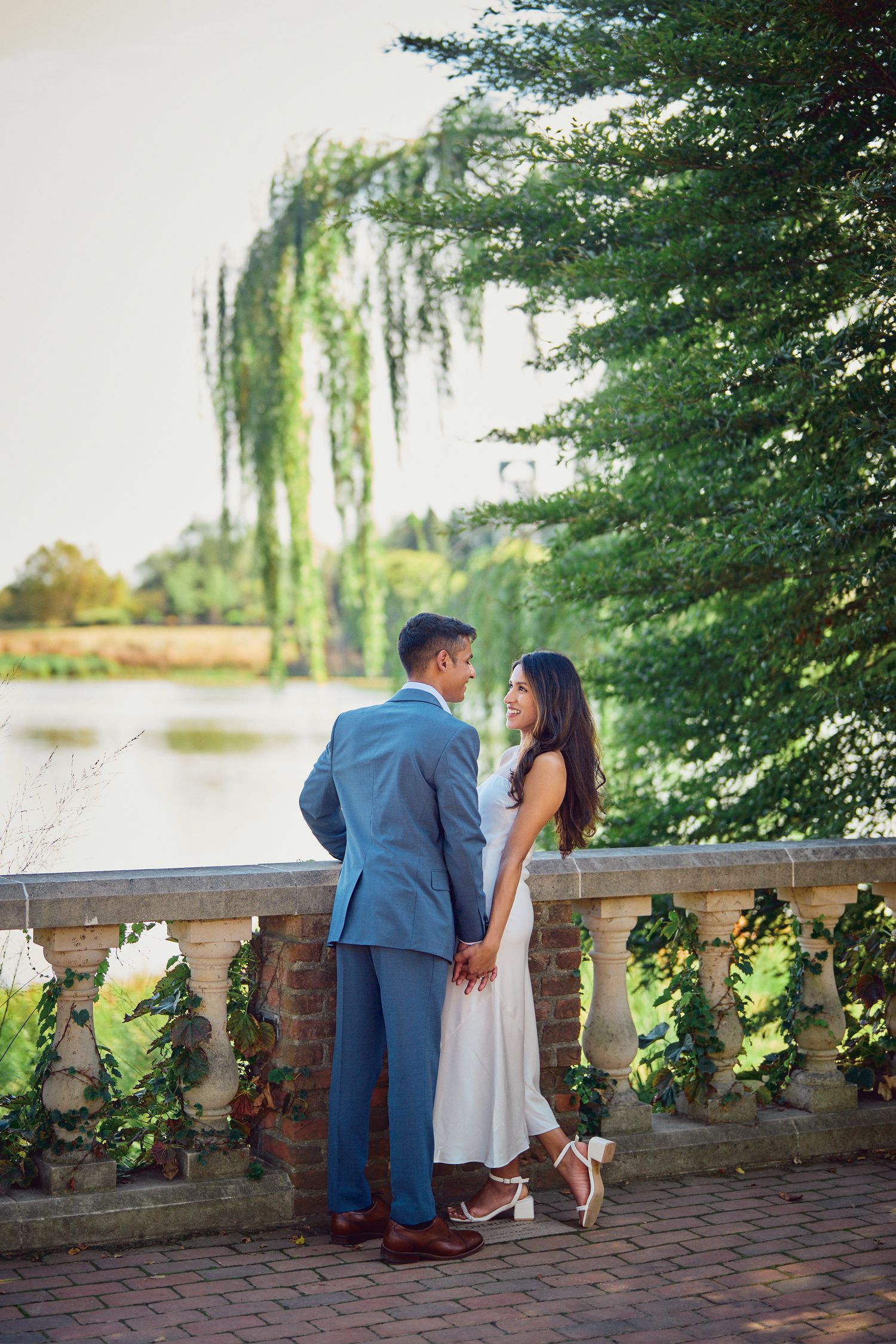 Couple in formal attire standing on brick terrace with willow tree and pond in romantic garden setting.