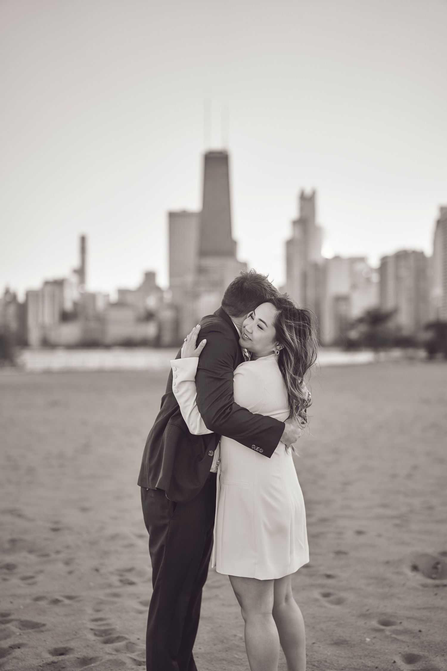 A romantic couple embraces on a beach with the Chicago skyline visible in the background in black and white.