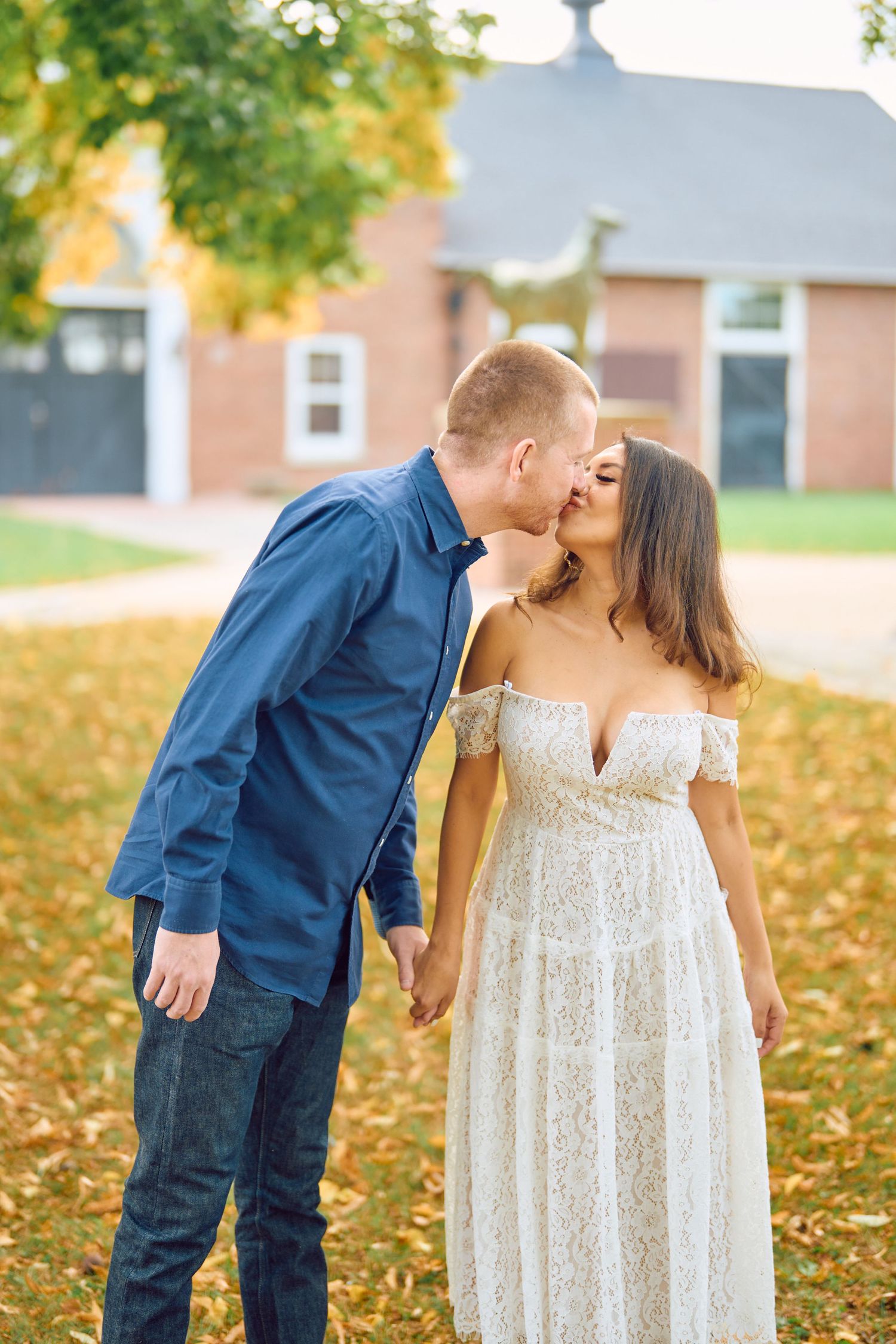 A couple shares a romantic kiss on fallen autumn leaves while standing in front of a brick building.