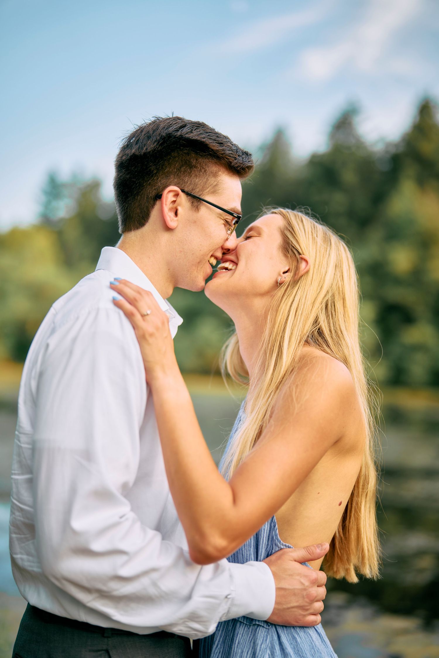 A romantic couple shares a tender embrace outdoors on a sunny day with trees and a lake in the background.