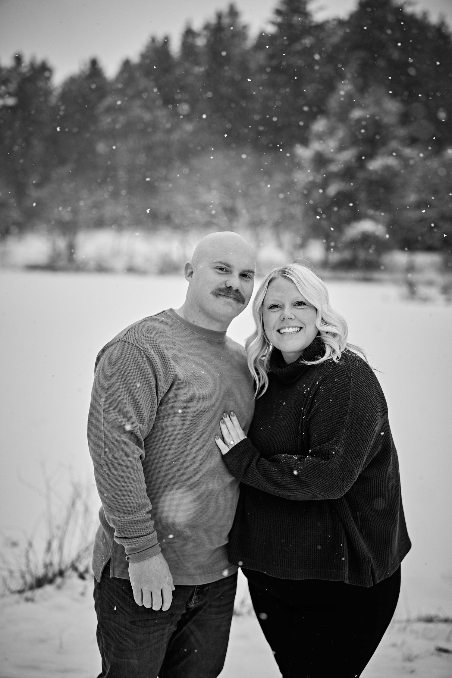 A couple poses for a black and white winter portrait with snow falling in a forest landscape.