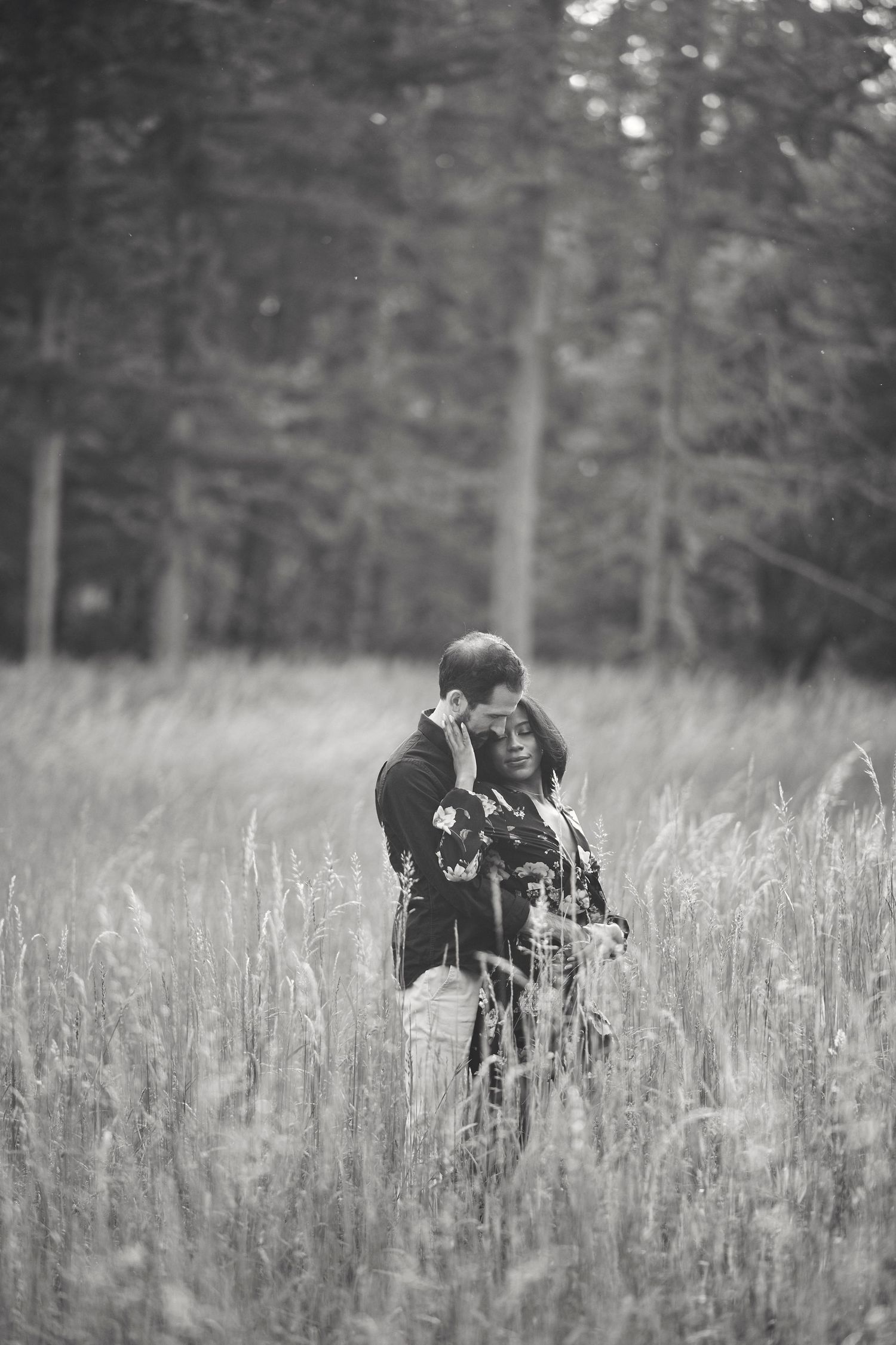 Romantic black and white photo of a couple embracing in a tall grass meadow with forest in the background.