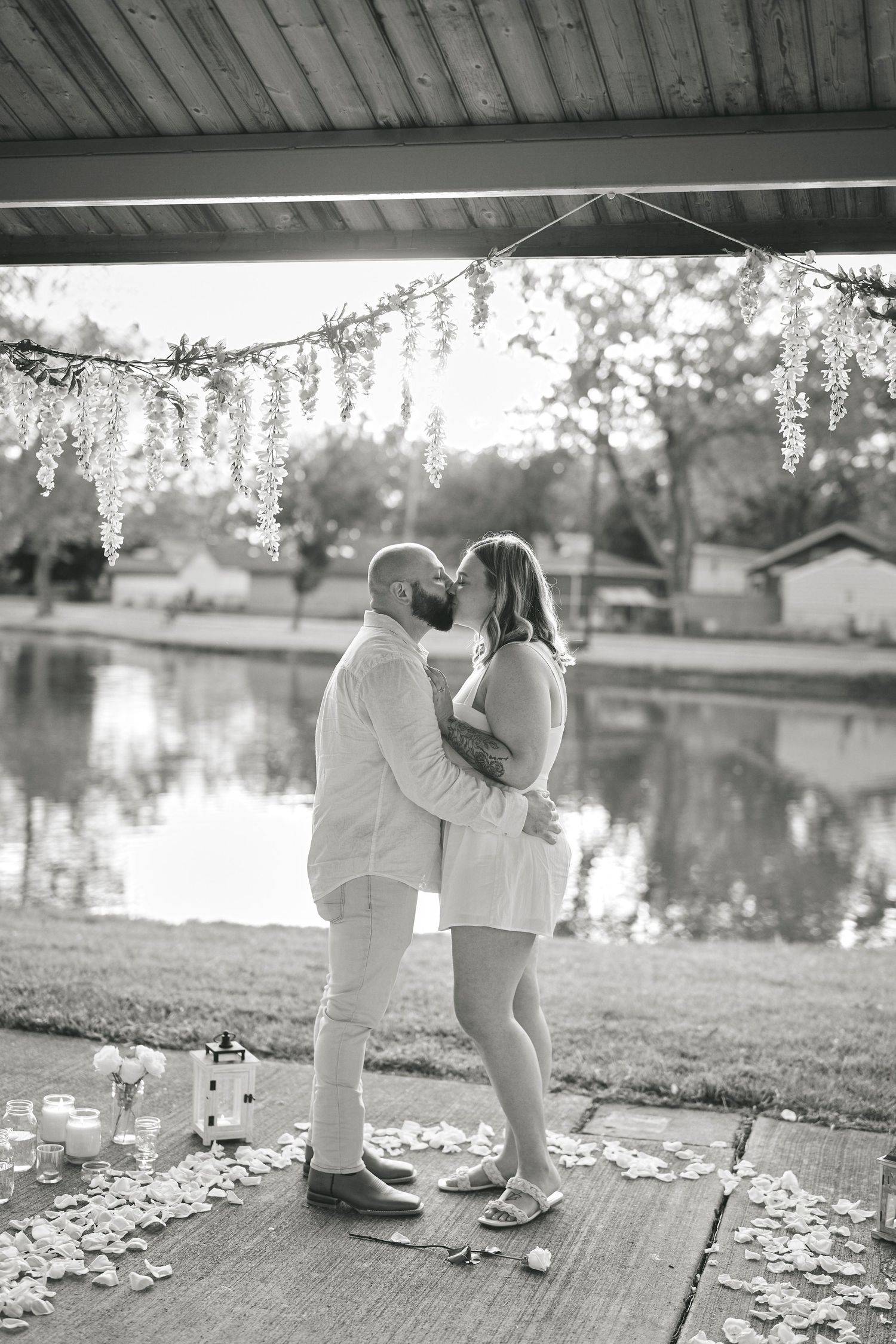 A romantic couple shares a tender moment by a lake under decorative lights on a wooden deck with scattered flower petals.