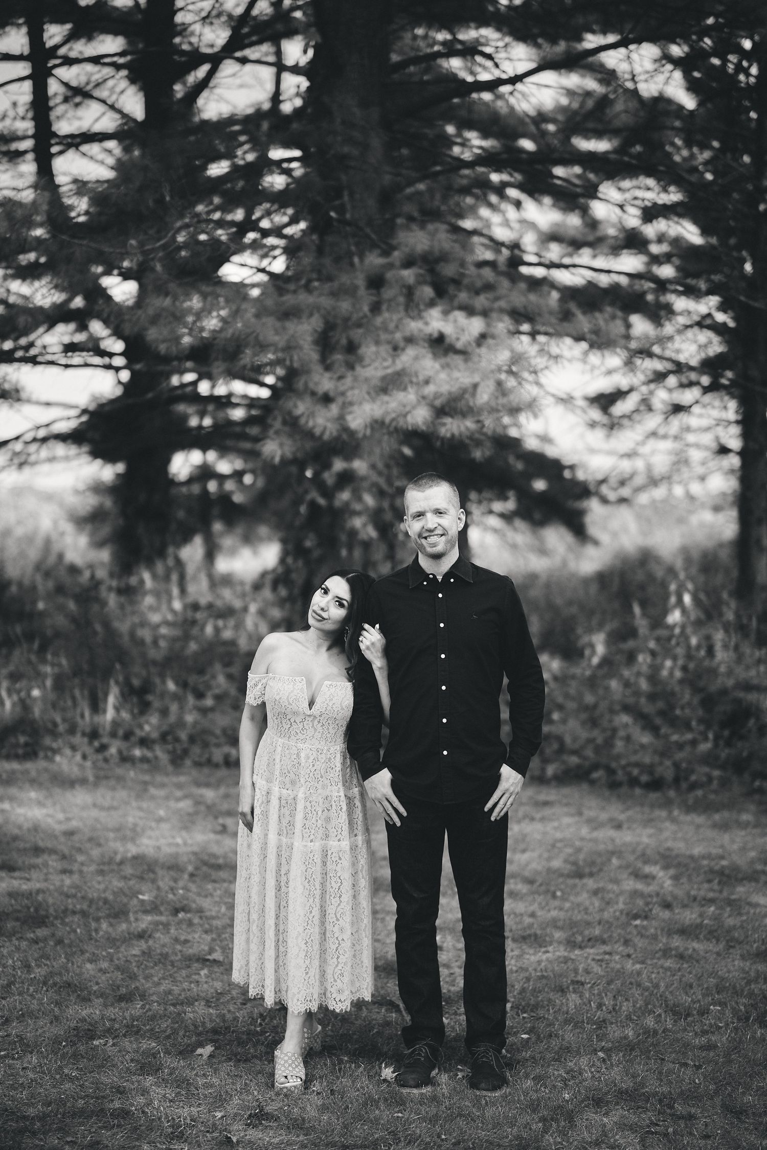 A black and white photograph of a couple standing together outdoors in a wooded setting.