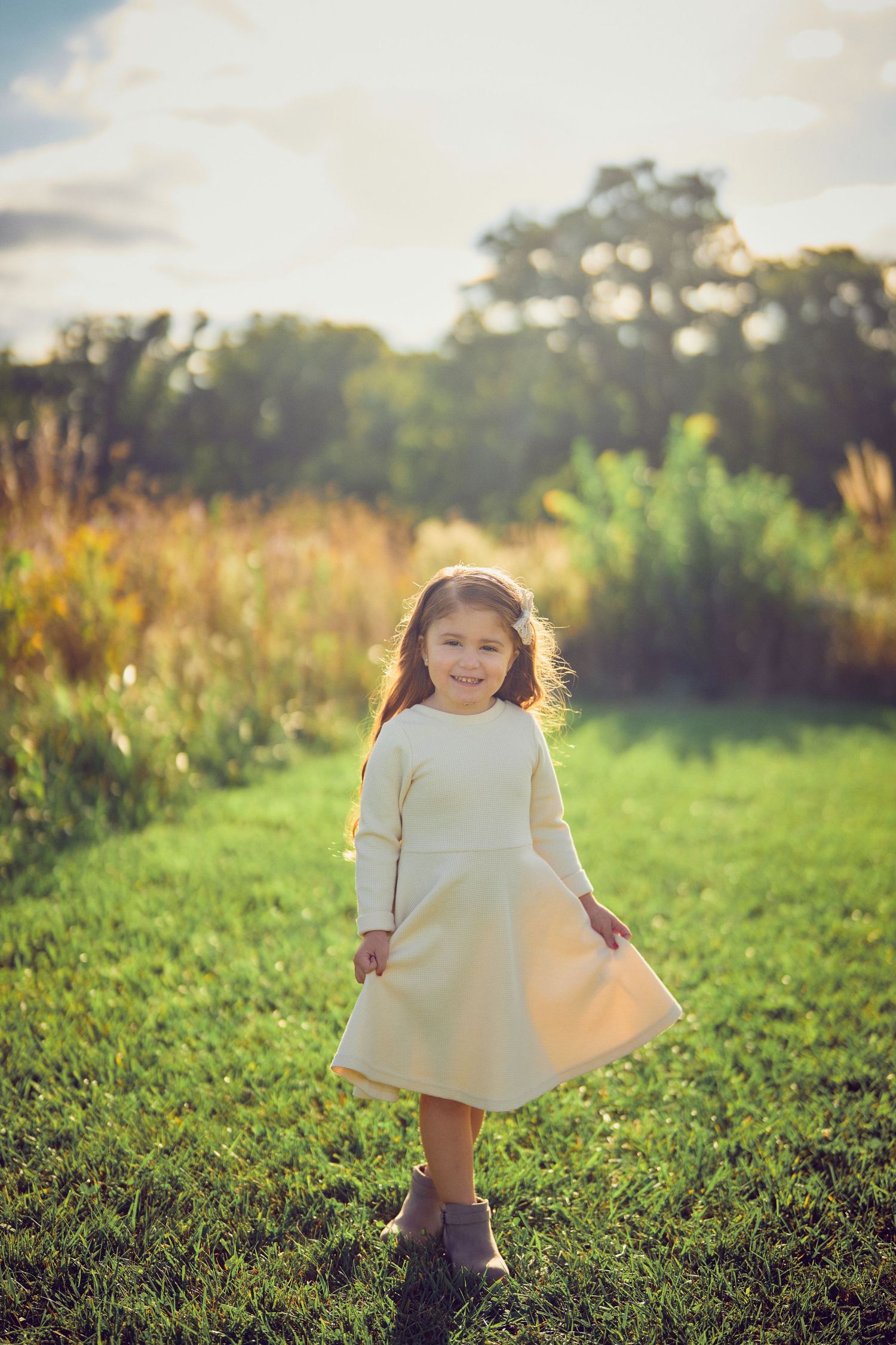A young child in a cream-colored dress twirls playfully in a sunlit meadow at golden hour.