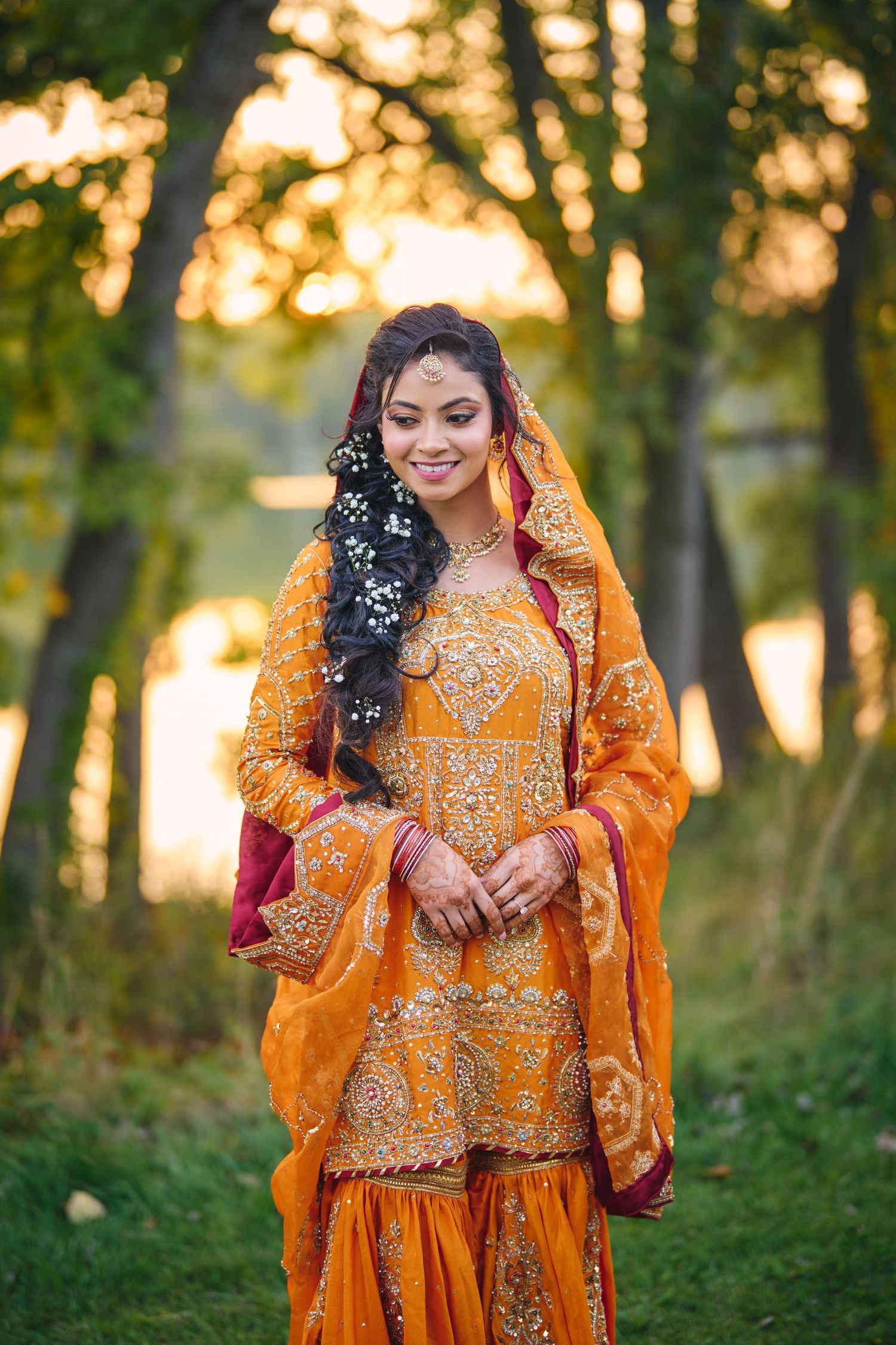 Traditional South Asian bride in orange embroidered dress and veil poses in forest during golden hour sunset.