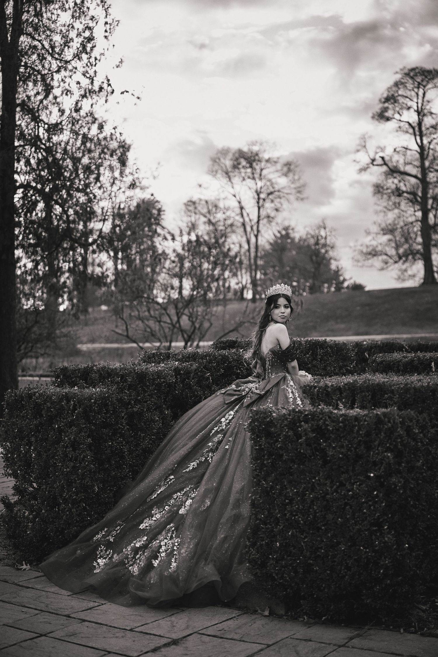 A figure in a flowing gown poses dramatically against a garden hedge in a black and white artistic photograph series.