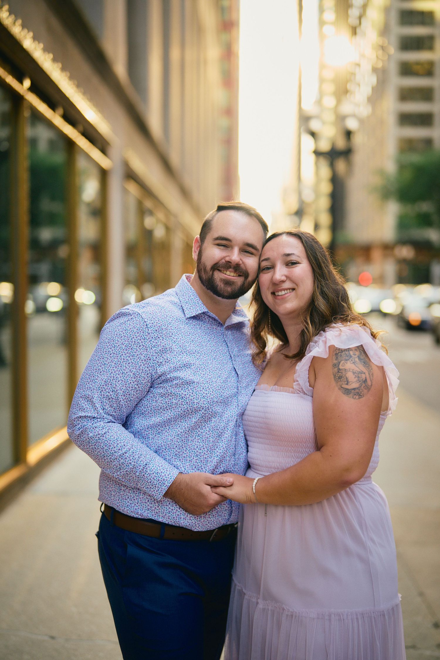 A couple in formal wear smiles and poses together on an illuminated urban sidewalk with storefronts behind them.