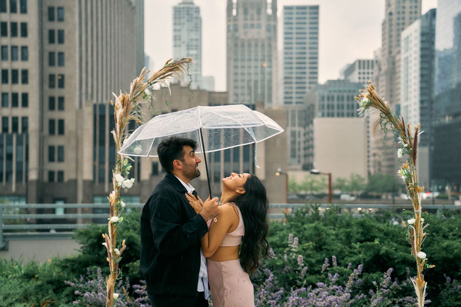A couple in formal wear shares an intimate moment under a clear umbrella against a cityscape backdrop on a rooftop garden.