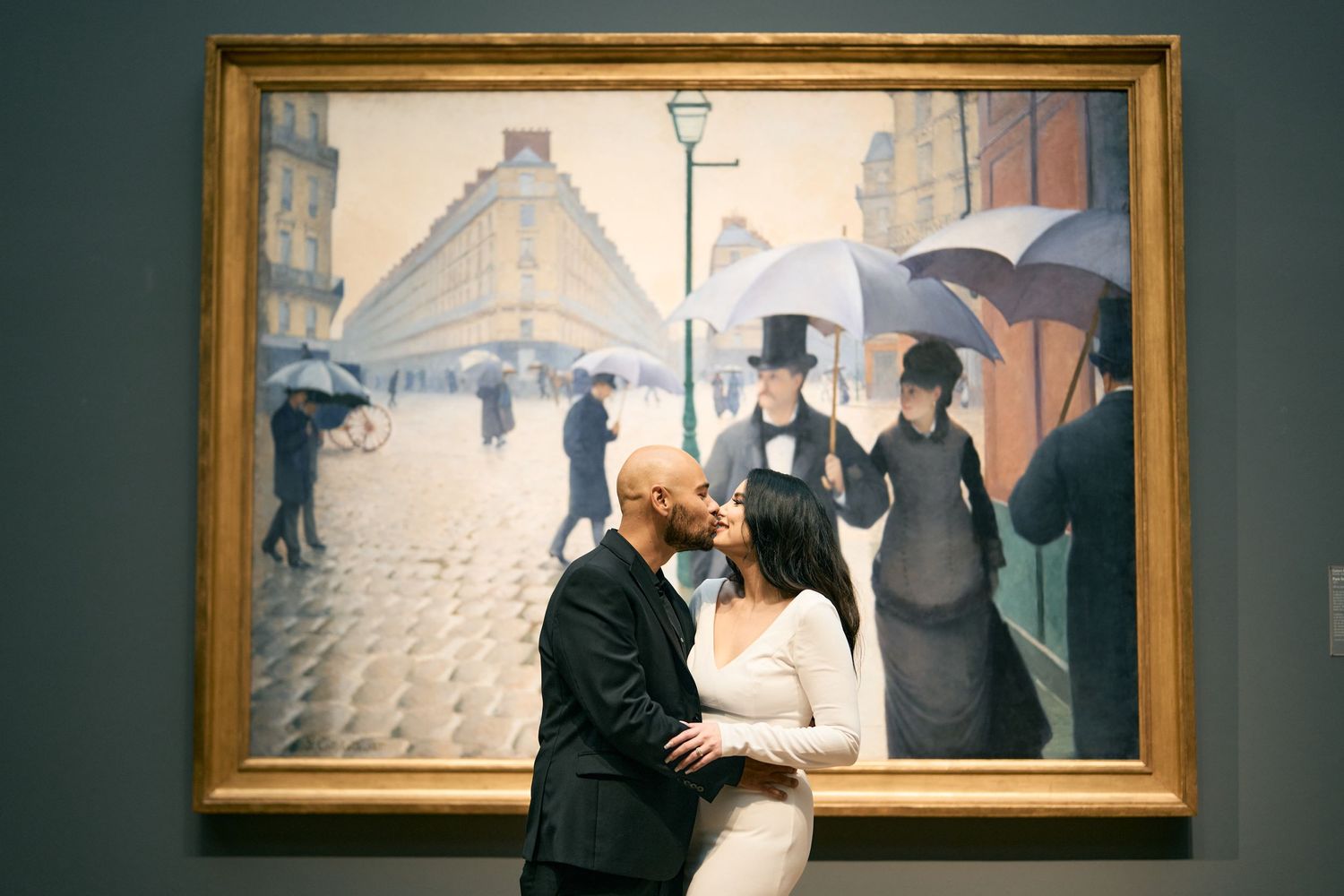 Visitors view and mirror a famous rainy Paris street scene painting displayed in gold frame at an art gallery.