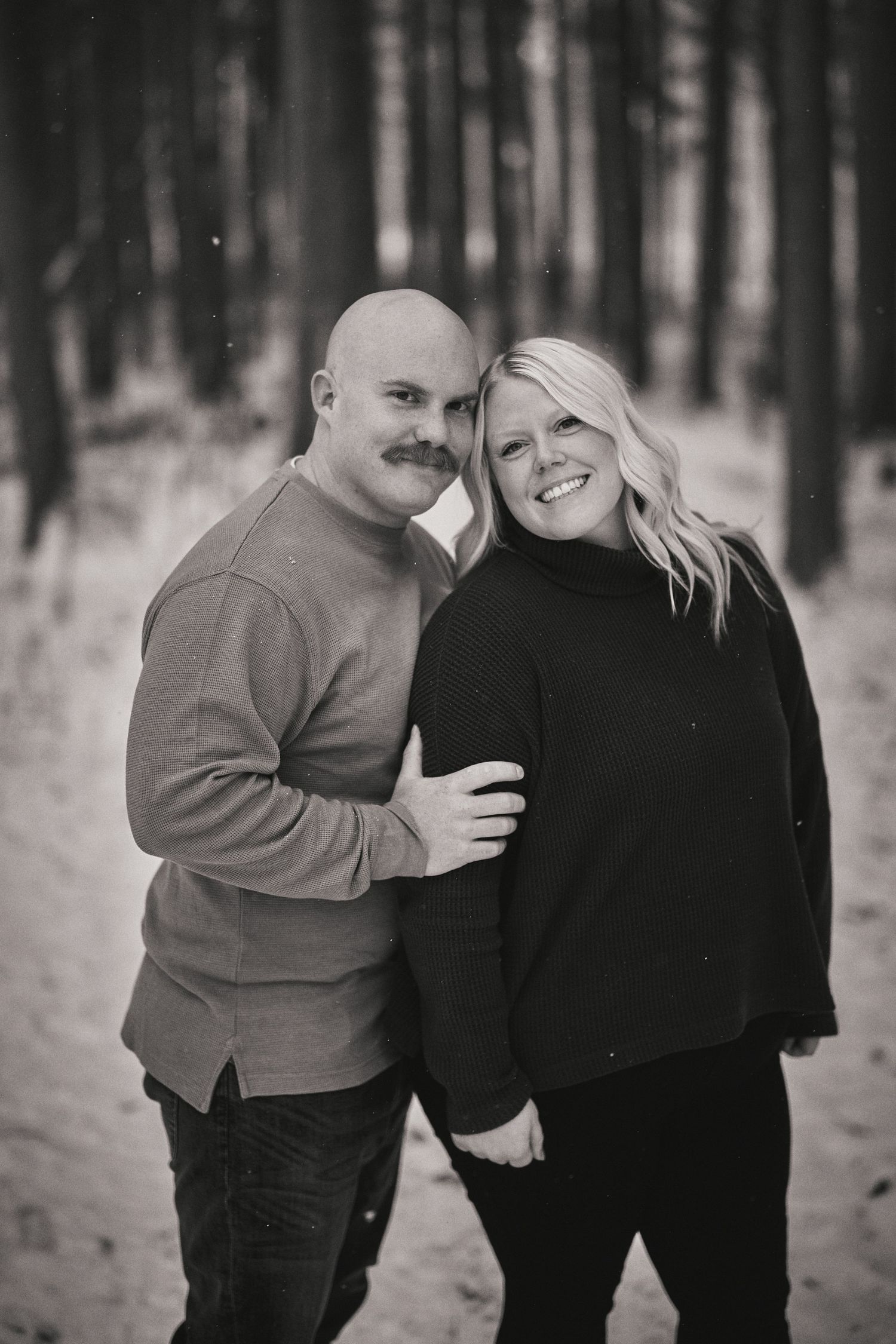 Black and white portrait of a couple embracing and smiling while standing in a forest setting.