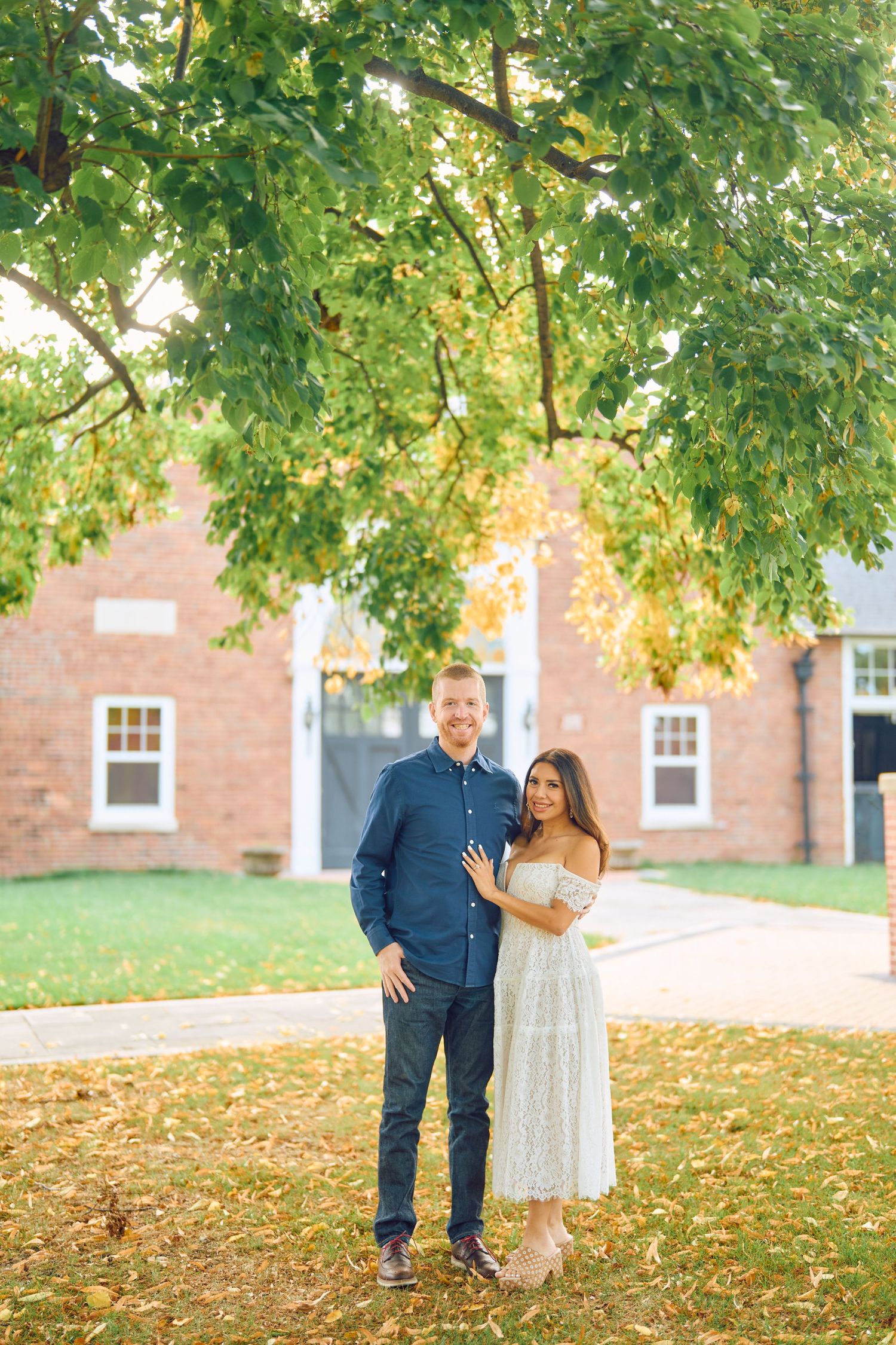 A couple poses under a leafy green tree in front of a brick building on a sunny autumn day.