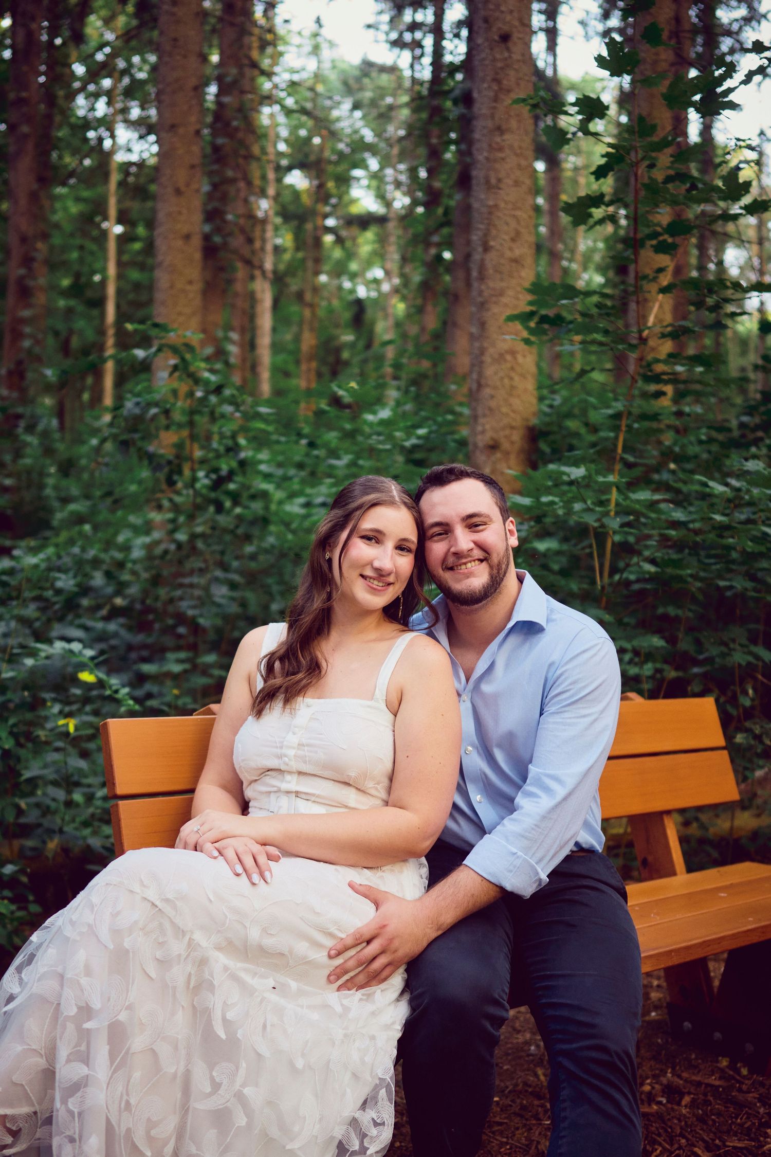 A beautiful outdoor engagement photo showing a couple sitting on a wooden bench surrounded by tall pine trees.