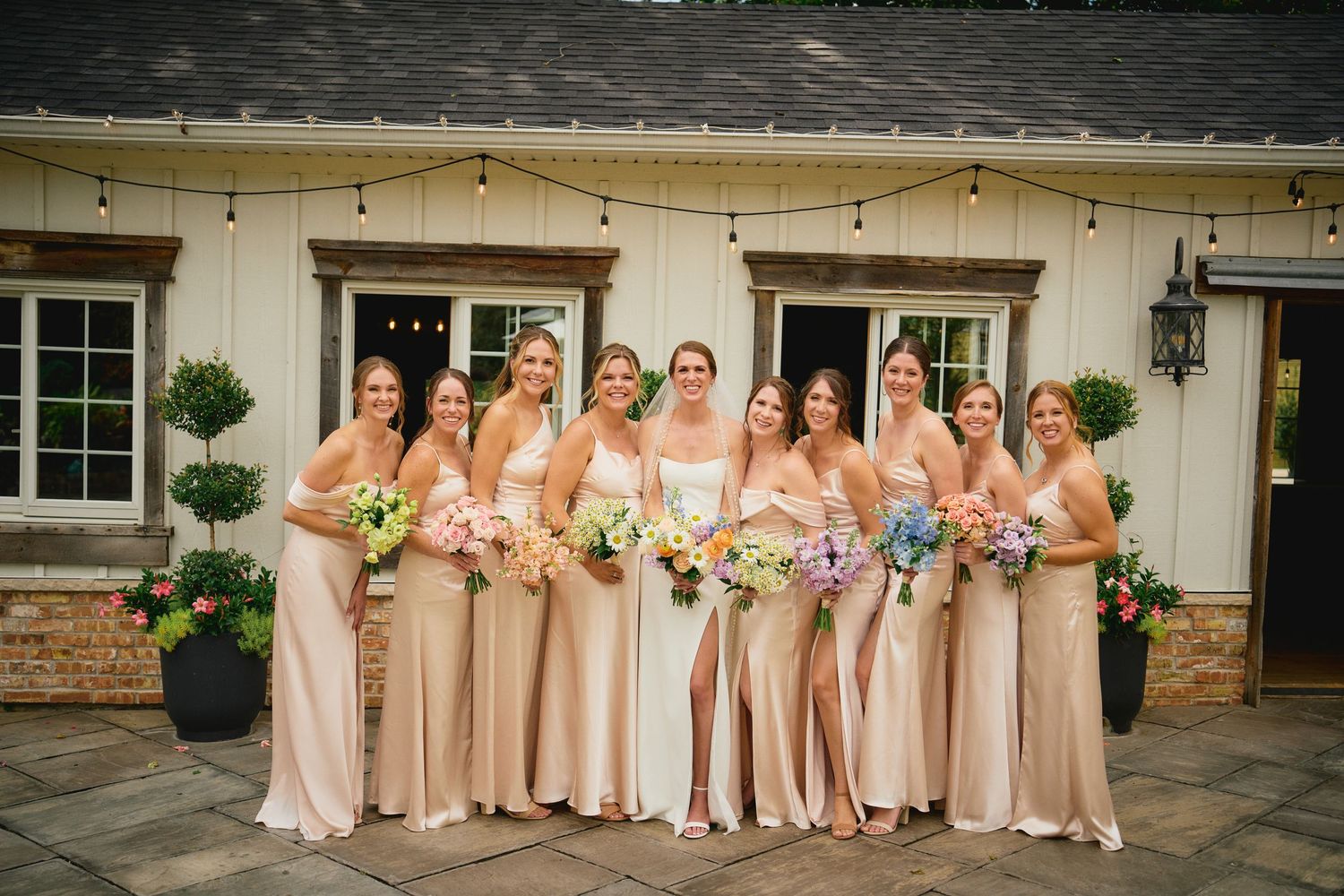 Elegant bridal party in blush pink floor-length dresses poses together holding colorful bouquets on a stone patio.
