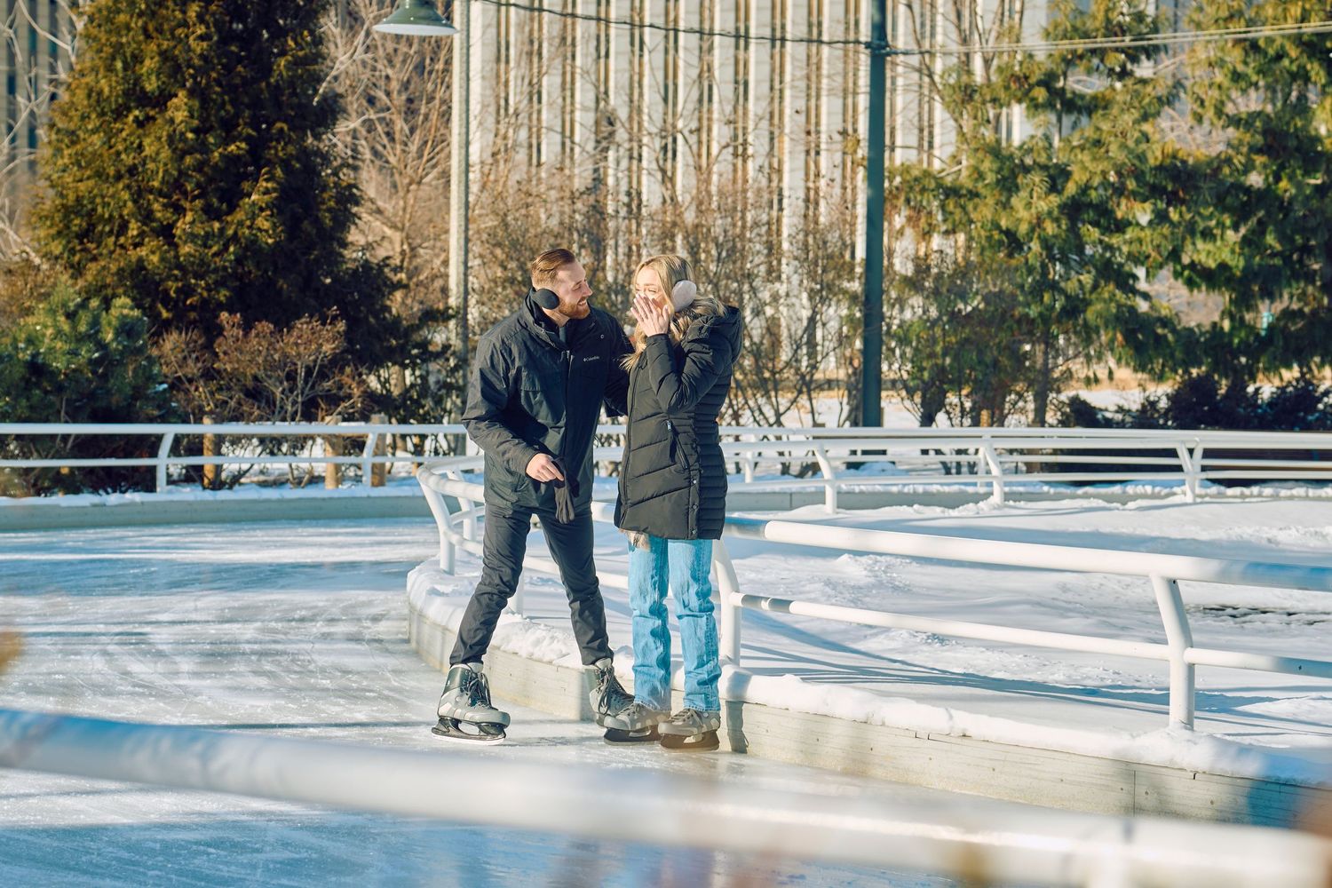 Two people enjoying ice skating together on an outdoor rink surrounded by trees on a sunny winter day.