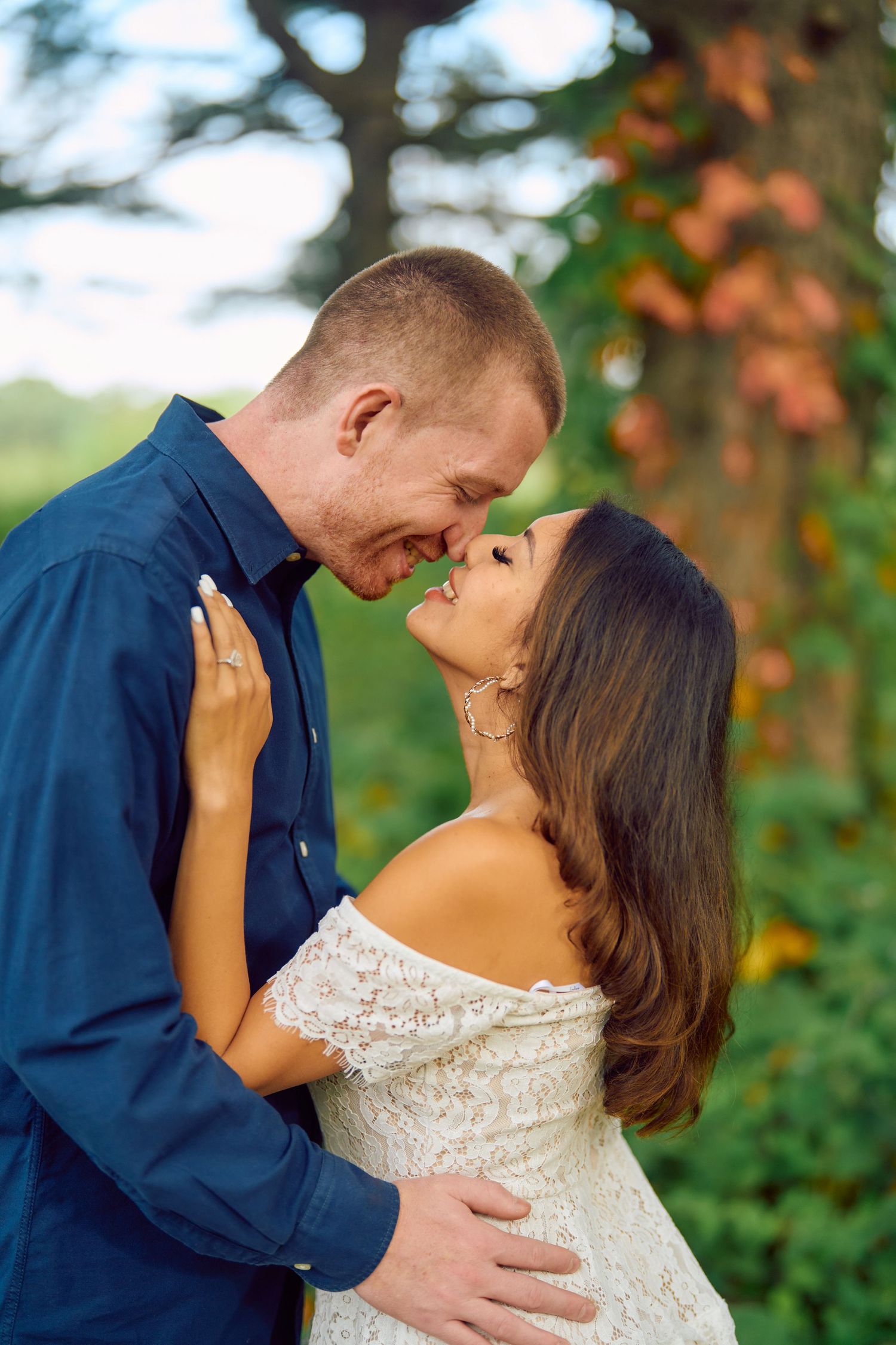 A romantic couple embraces outdoors, with her wearing a white lace off-shoulder dress and him in a navy button-down shirt.
