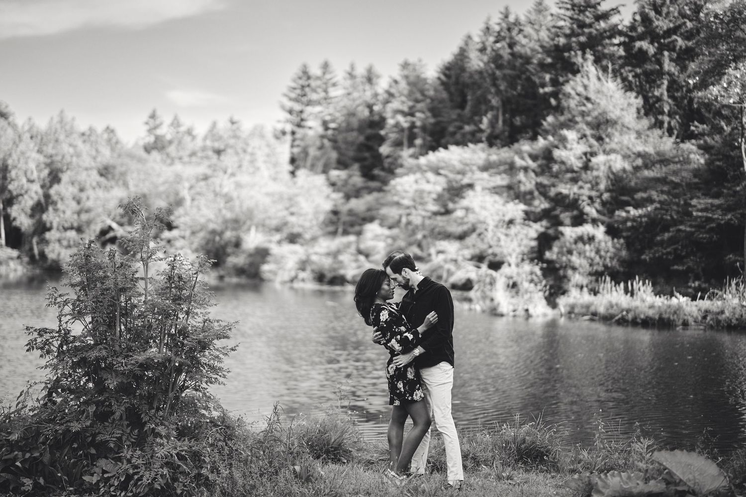 A romantic black and white photo of two people embracing by a serene lake surrounded by pine trees.
