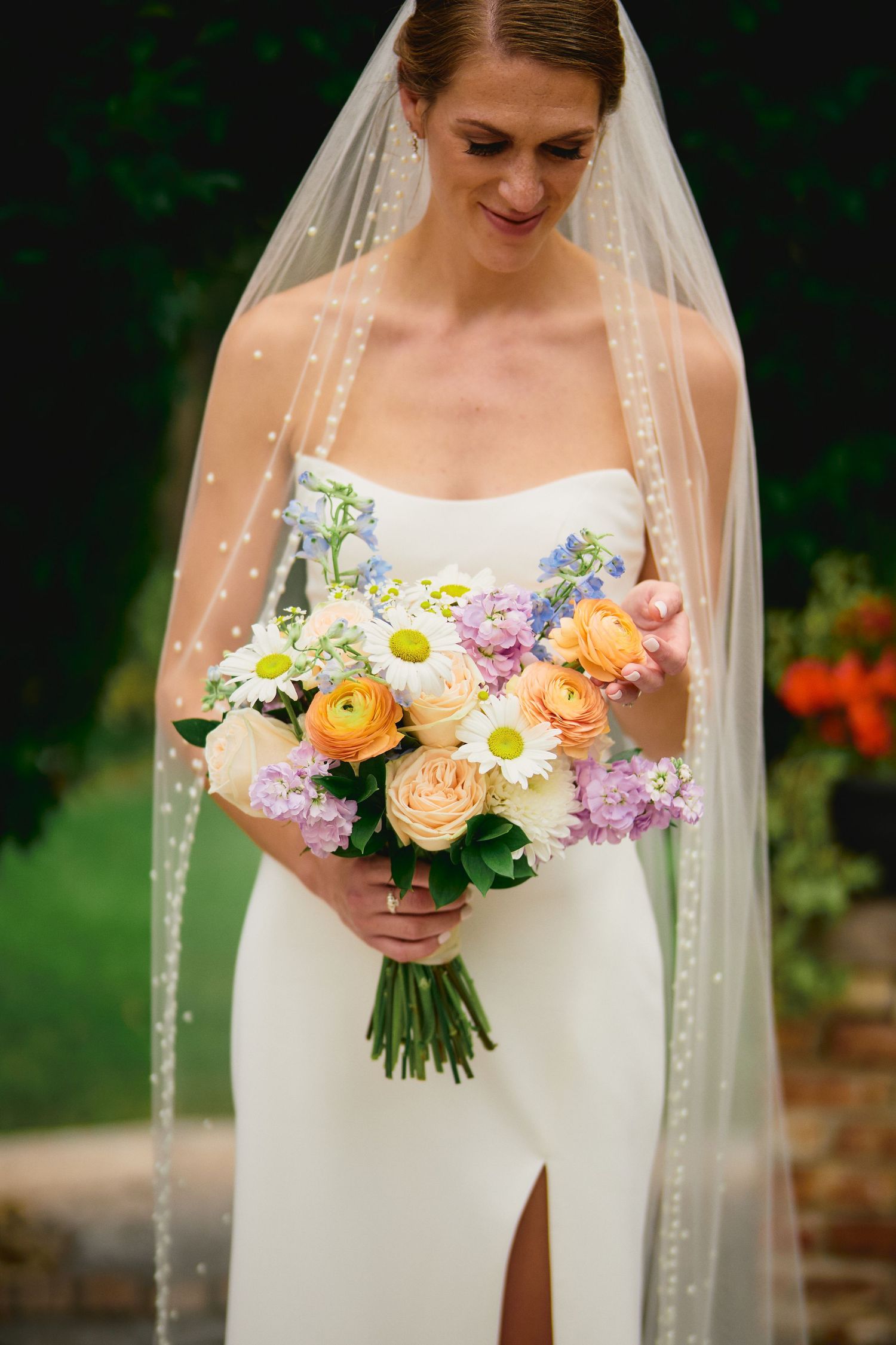 Beautiful bridal bouquet with orange ranunculus, white roses and purple lilac flowers paired with elegant beaded veil.