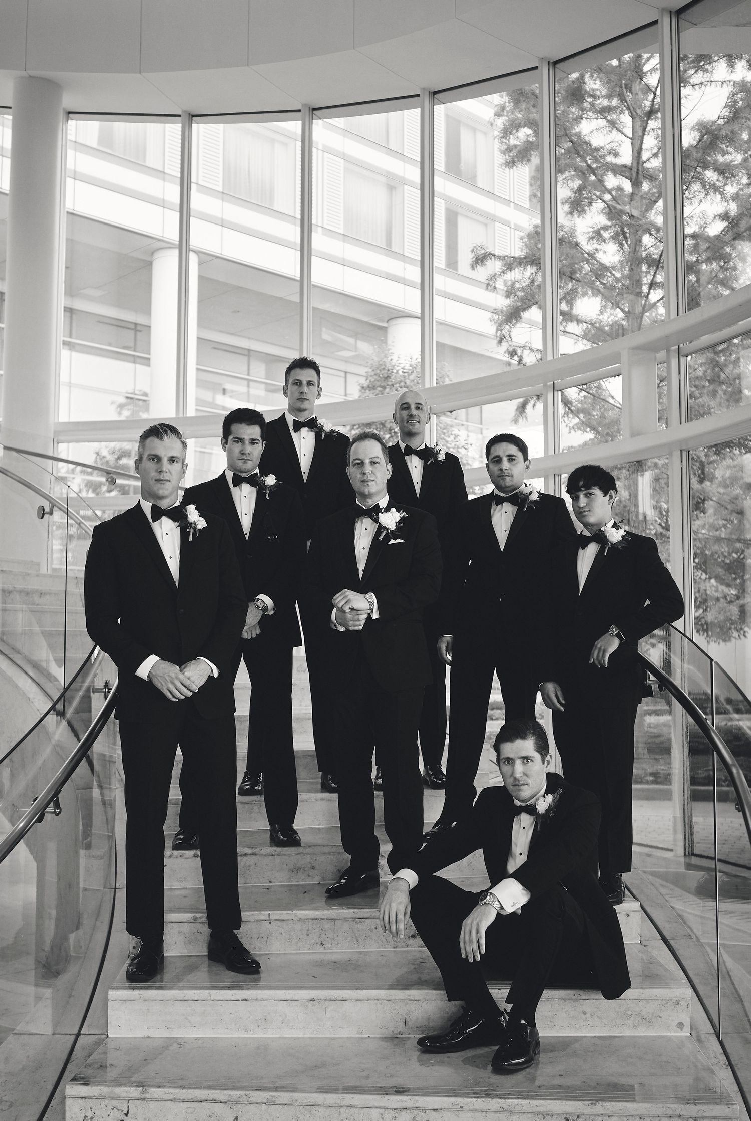 A black and white photograph of groomsmen in tuxedos posing on an elegant staircase with large windows.
