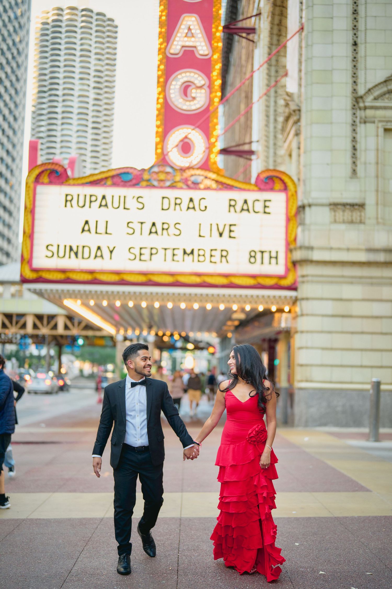 Couple holds hands walking beneath Chicago Theatre marquee advertising RuPaul's Drag Race All Stars show on State Street.