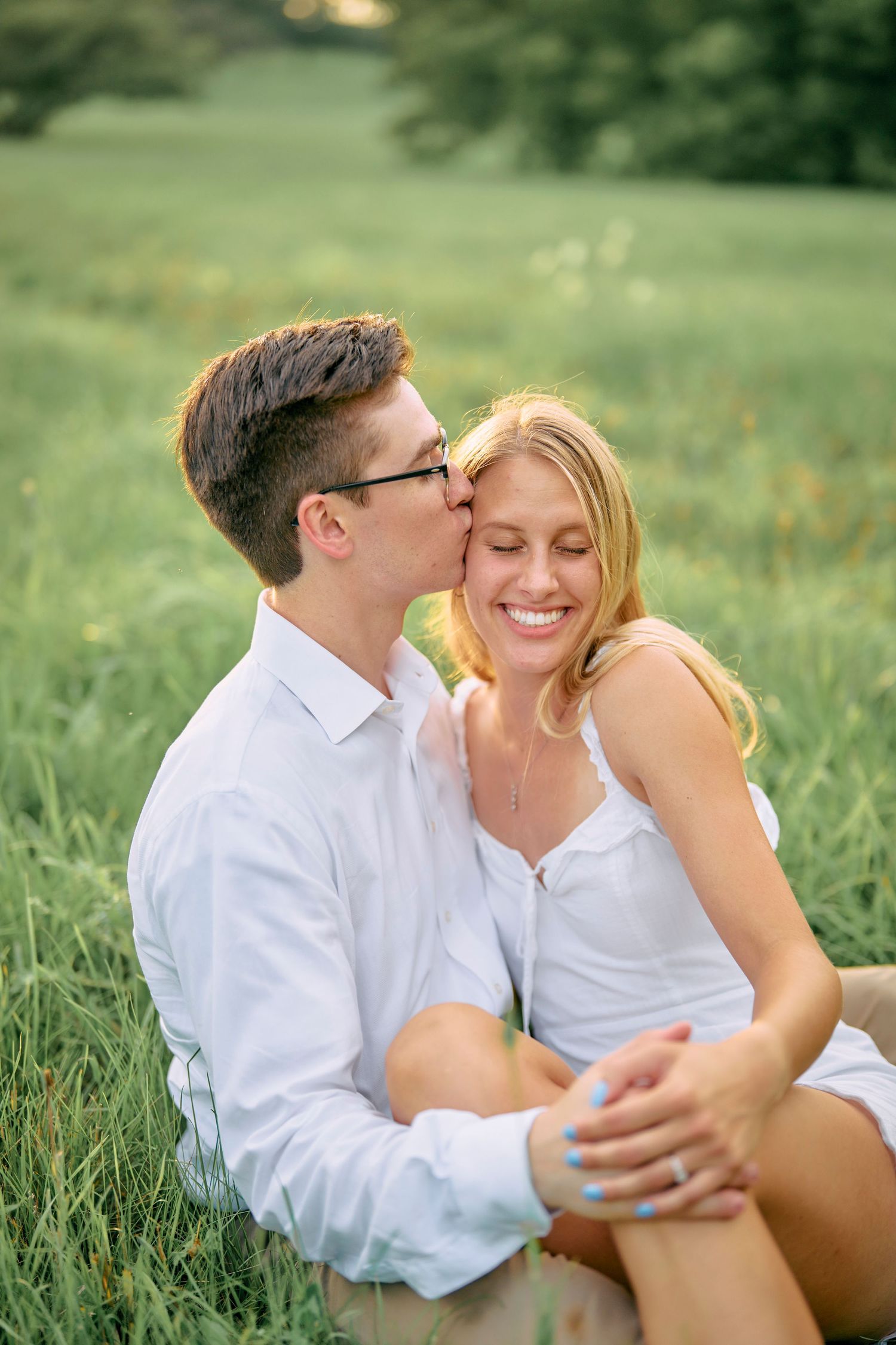 Two people in white outfits embrace lovingly while sitting in a sunlit grassy meadow on a warm summer day.