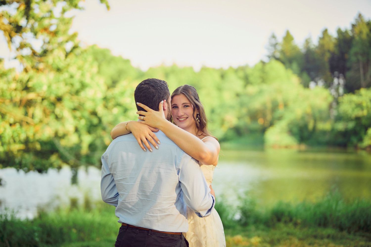 A couple embraces tenderly by a serene lake surrounded by lush green trees on a bright summer day.