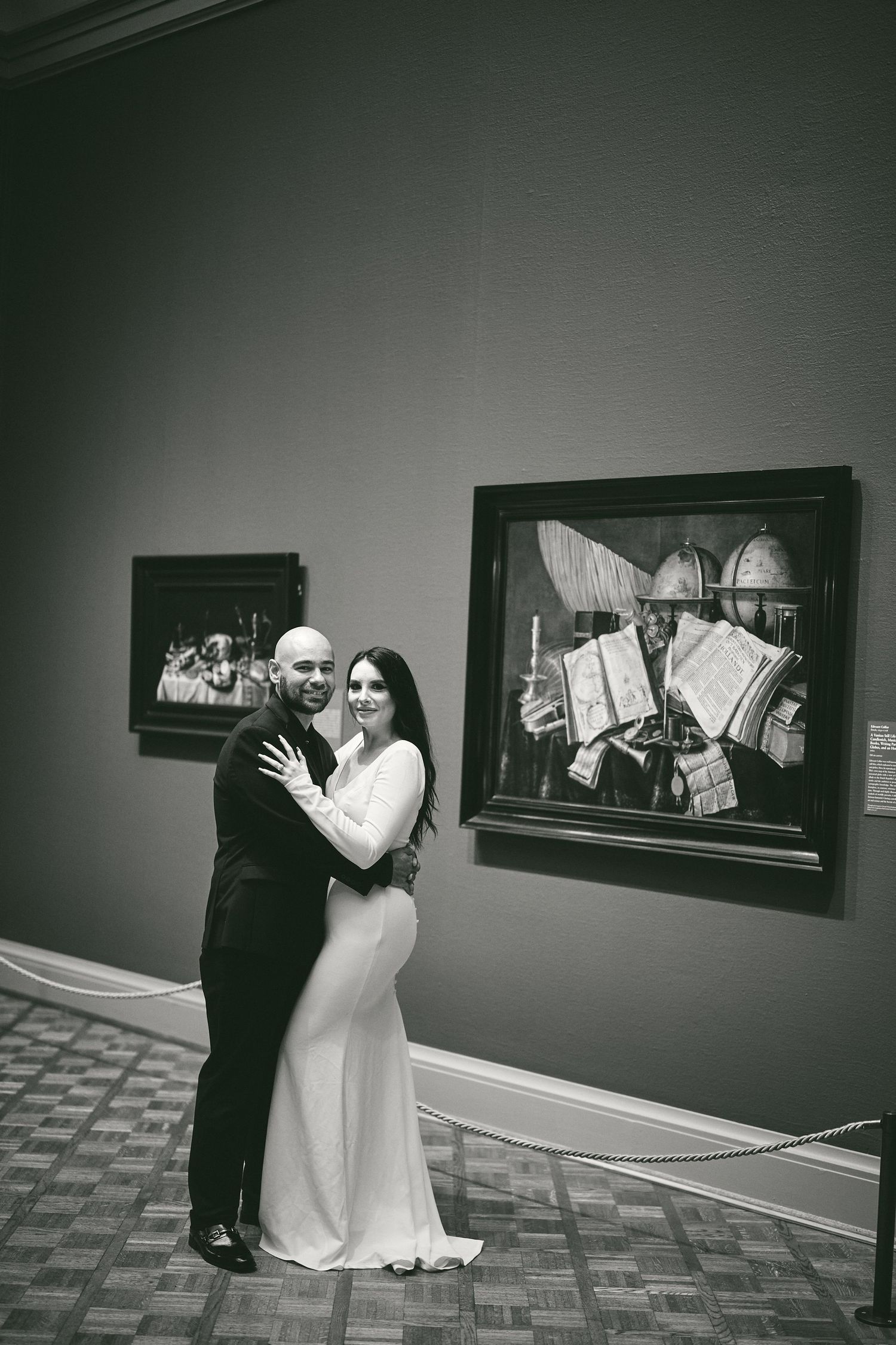 Black and white photo of a couple embracing in a museum gallery with framed artwork displayed on the wall.