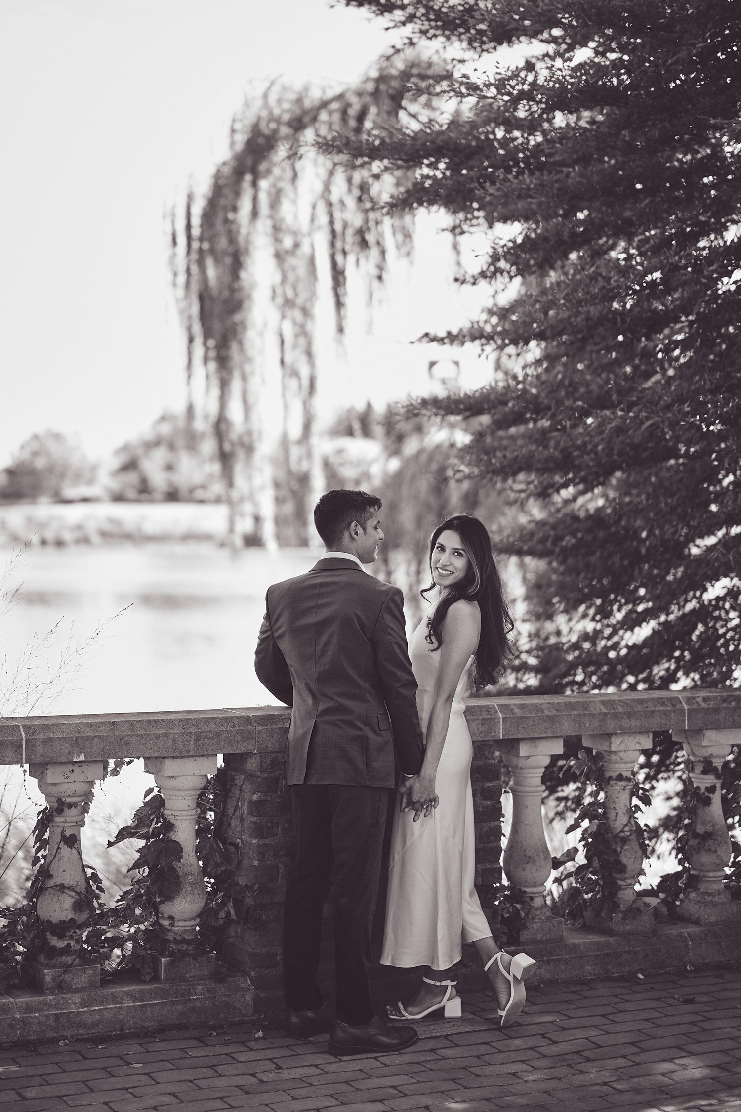 A couple in formal attire stands romantically on a stone balcony under a weeping willow tree in black and white.