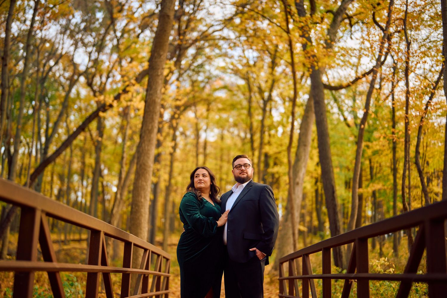 A couple stands on a rustic wooden bridge surrounded by golden autumn foliage in a sunlit forest.