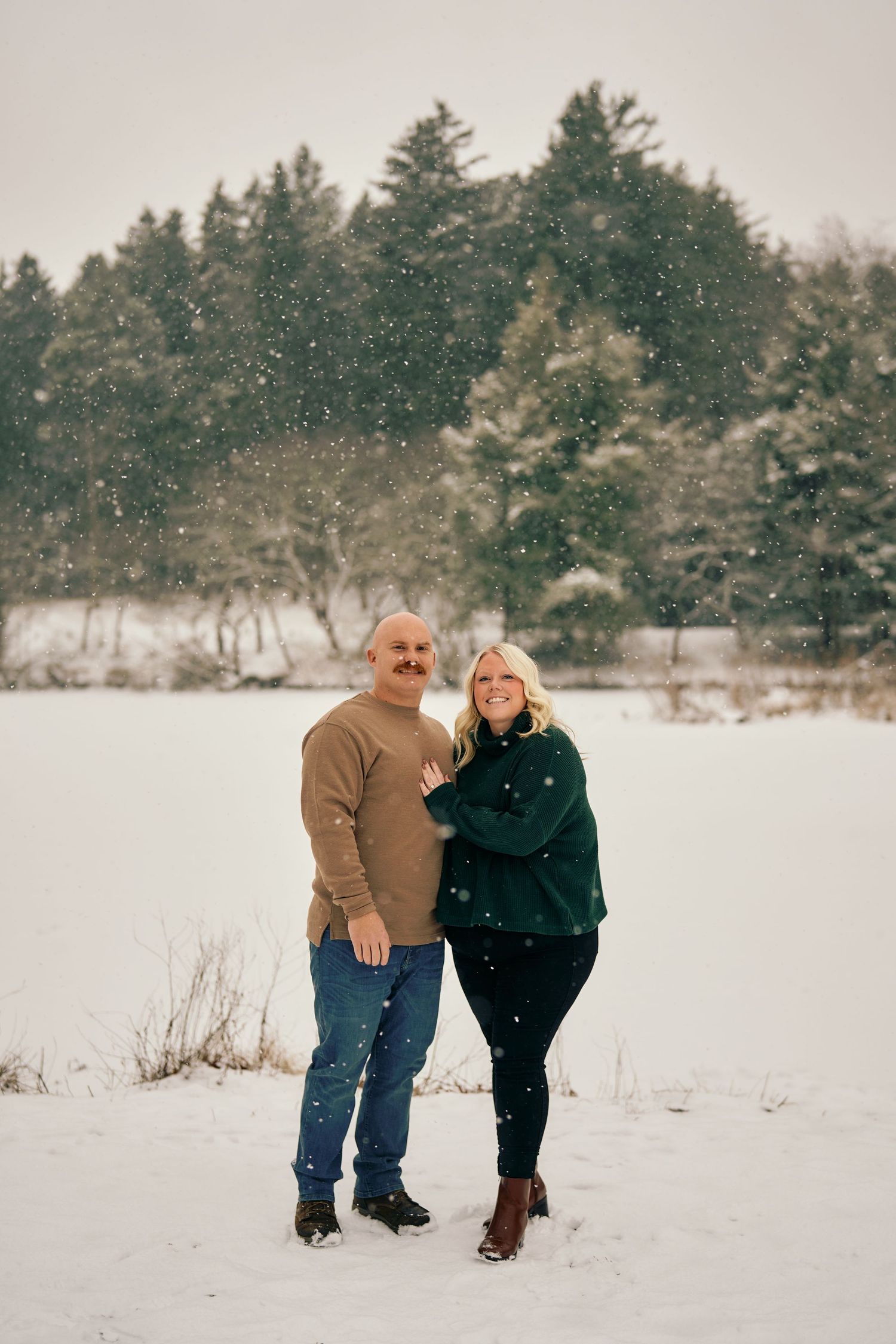A couple stands together in a snowy winter landscape with evergreen trees in the background during gentle snowfall.