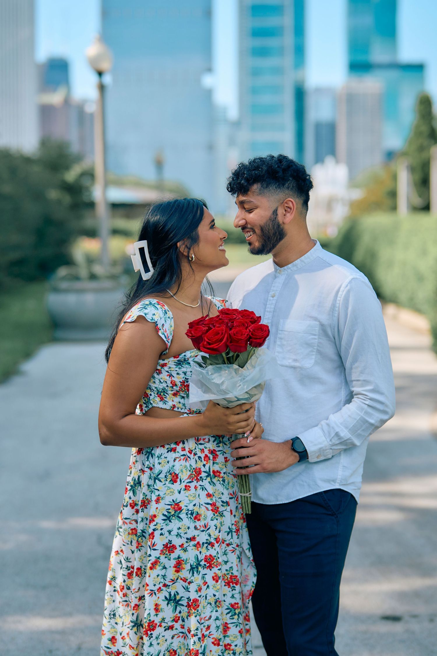 A couple shares a romantic moment in a city park as they hold a bouquet of red roses together on a sunny day.