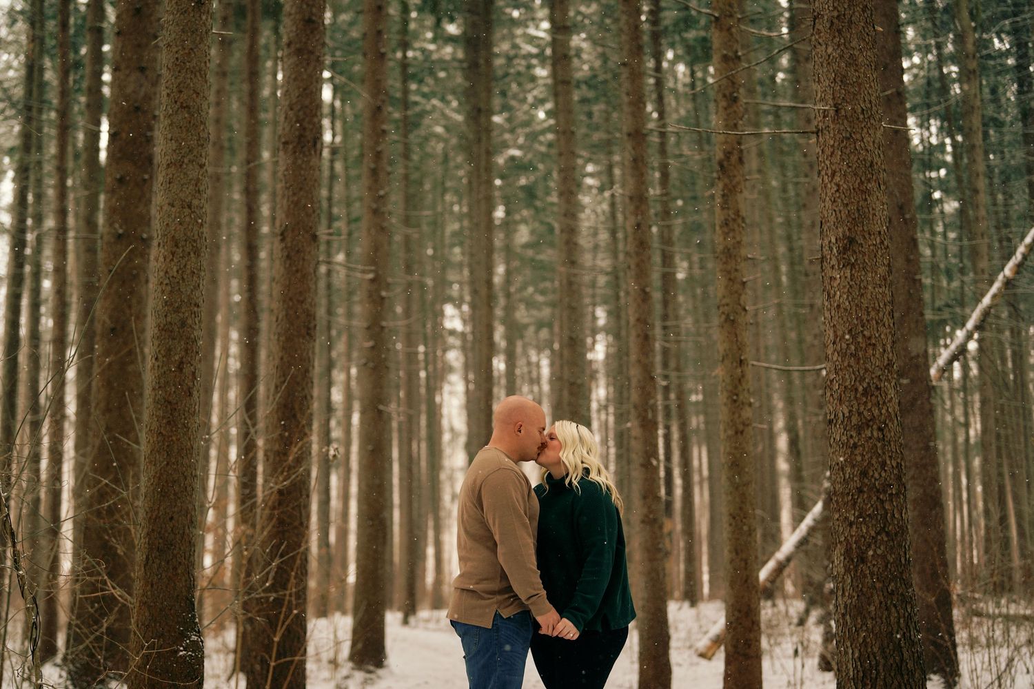 Couple in winter coats standing together among tall evergreen trees in snowy forest.