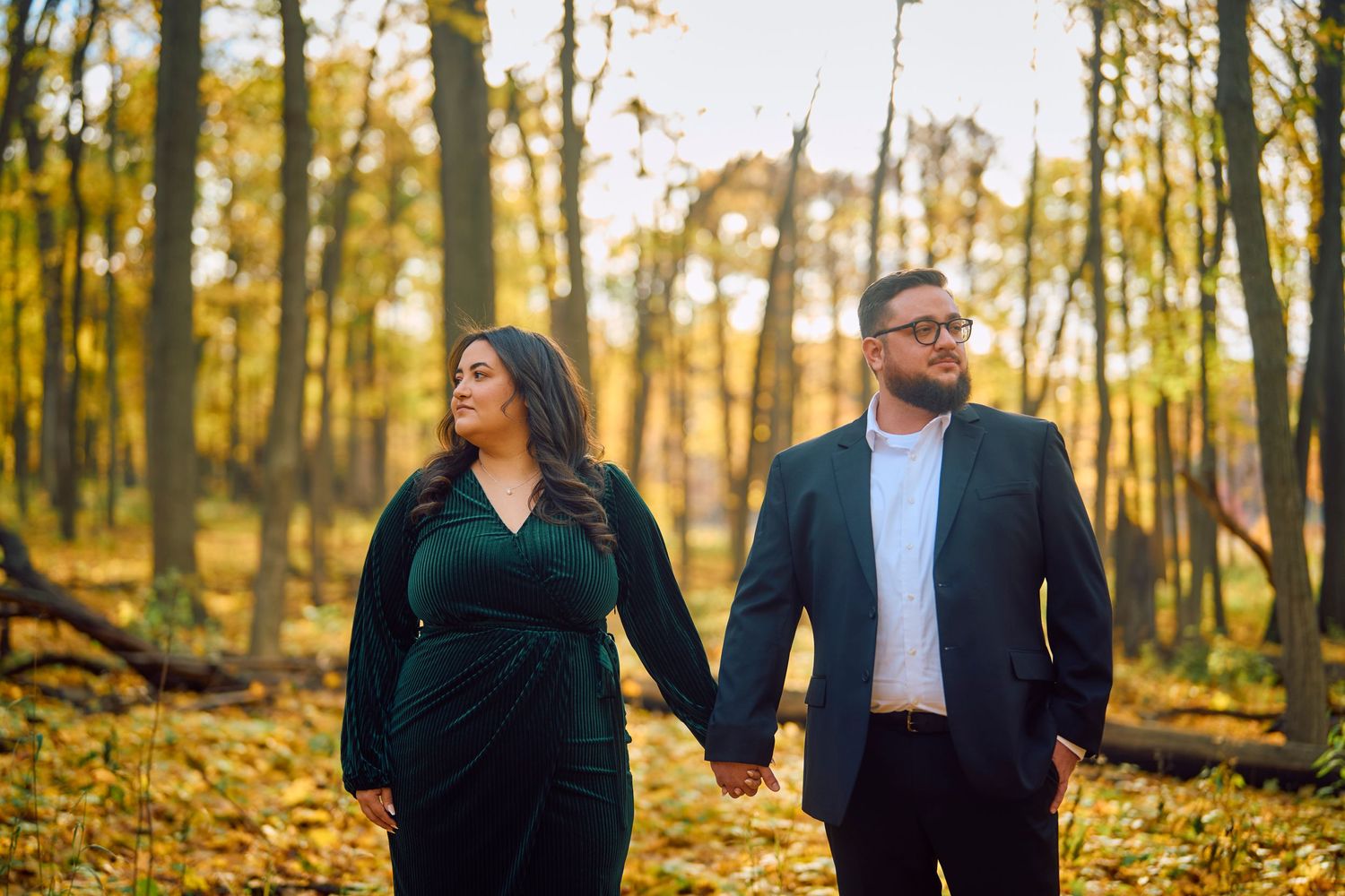 Two people walk hand in hand through a sunlit autumn forest with golden fallen leaves on the ground.