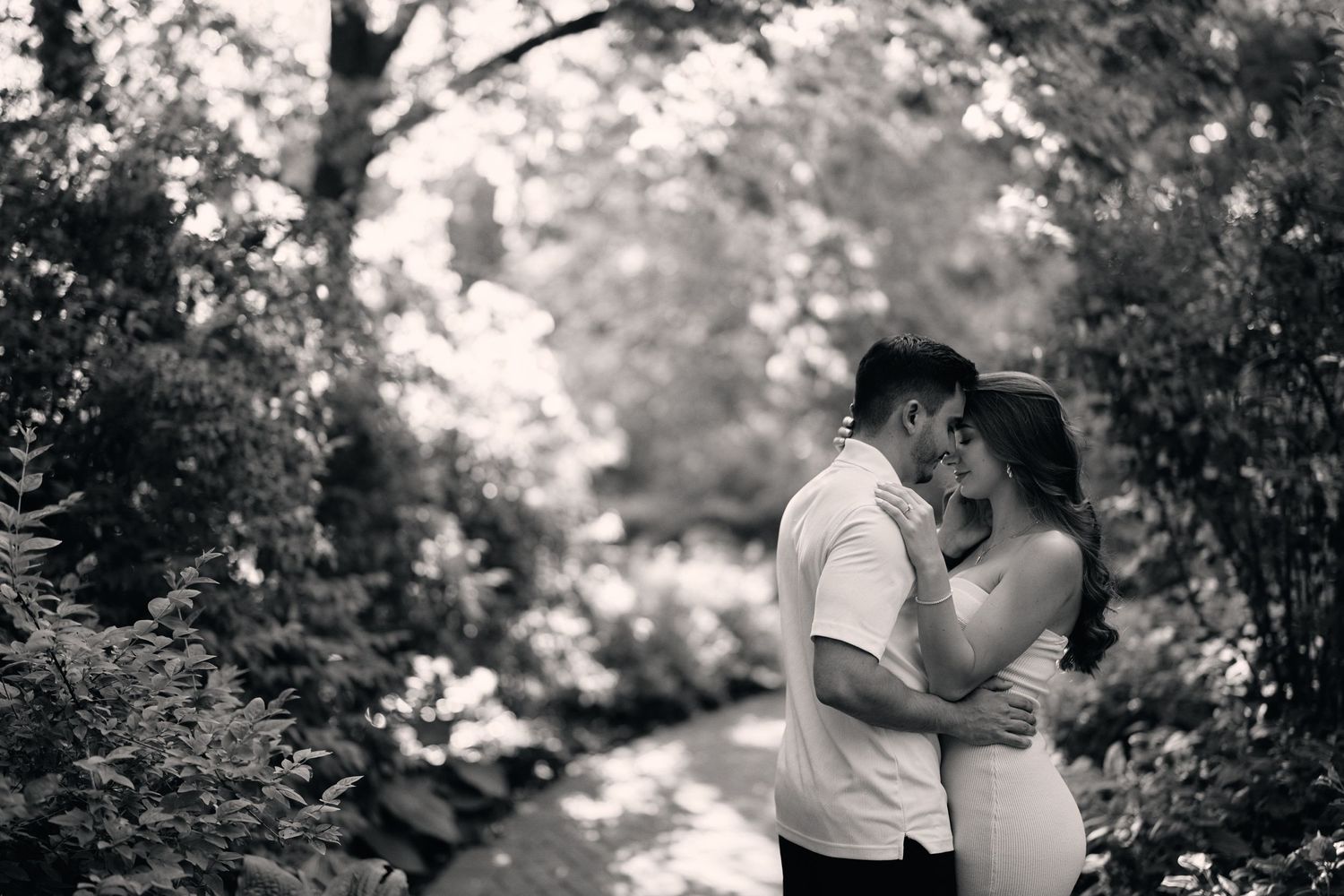 A romantic black and white maternity photo of a couple embracing on a tree-lined path in nature.