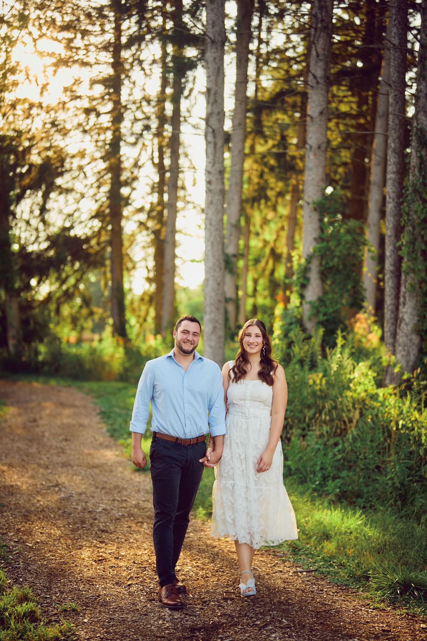 A couple walks hand in hand on a forest path during golden hour with sunlight filtering through tall trees.