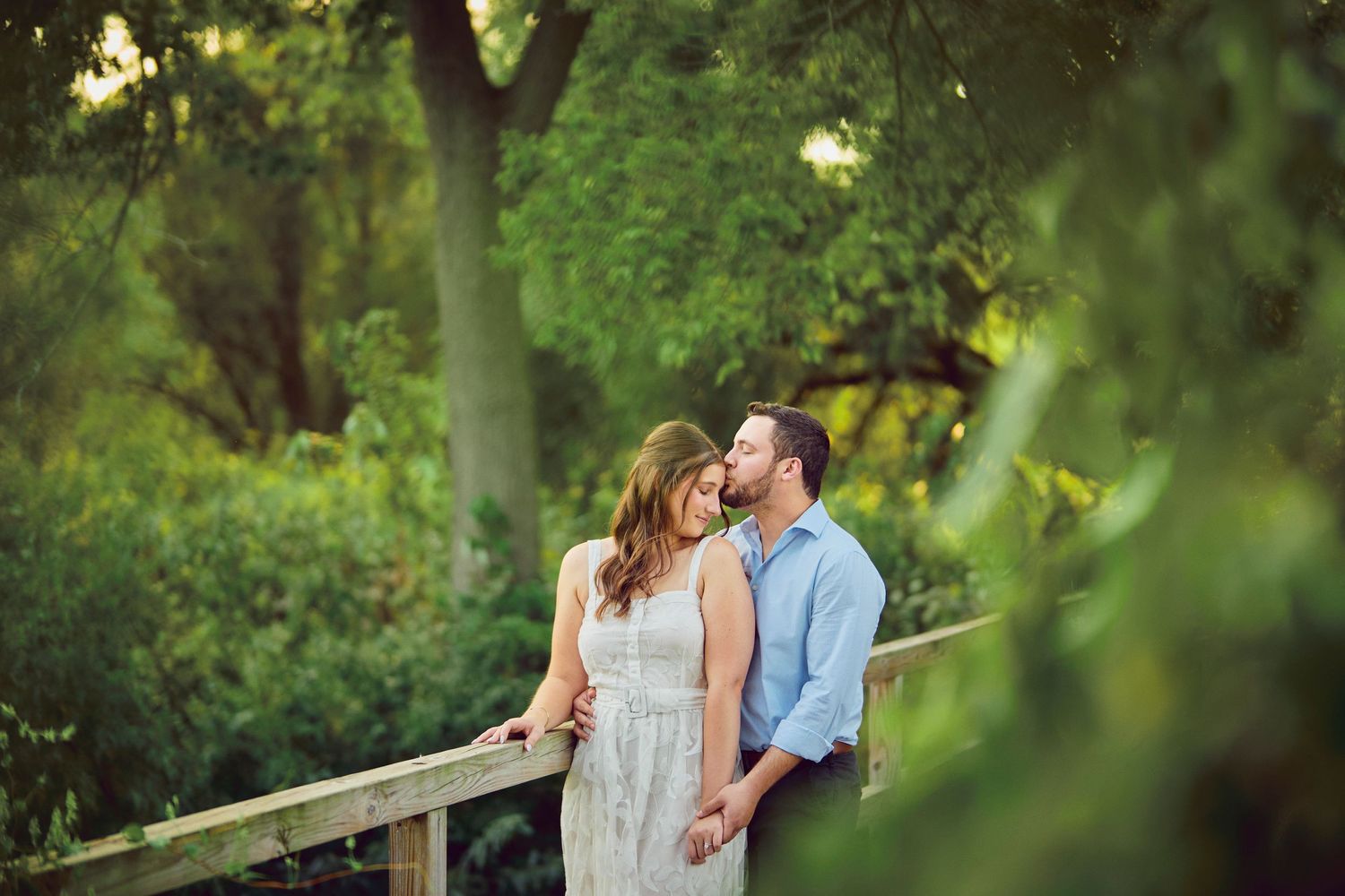 A romantic couple shares a tender moment on a wooden bridge surrounded by lush green foliage in a garden setting.