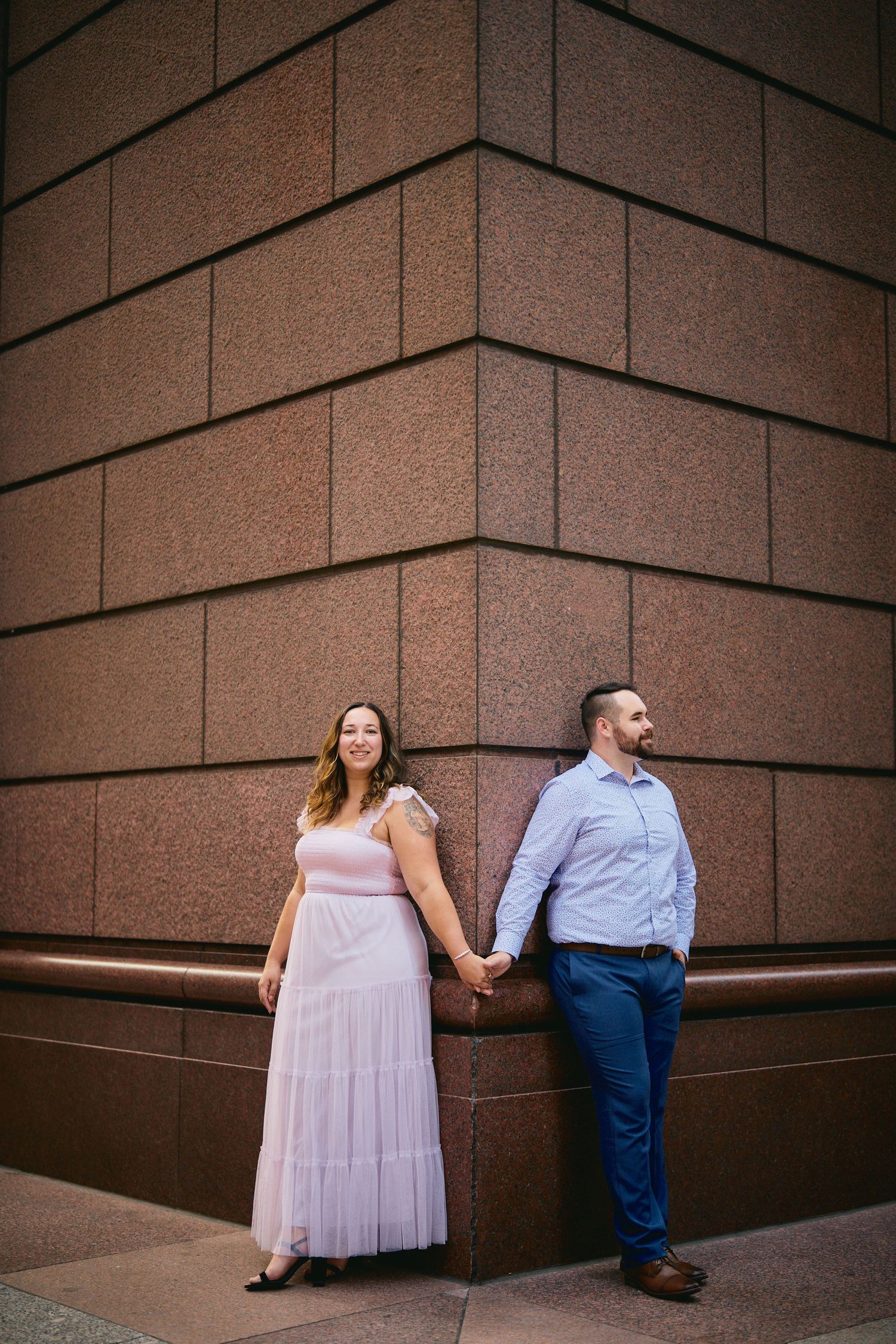 Couple holds hands while standing on opposite sides of a corner against a brown textured wall.