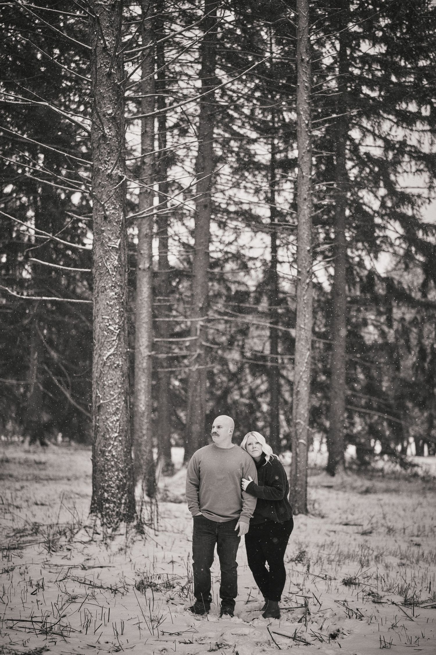 A black and white photo of a couple walking together through a snowy forest with tall pine trees.