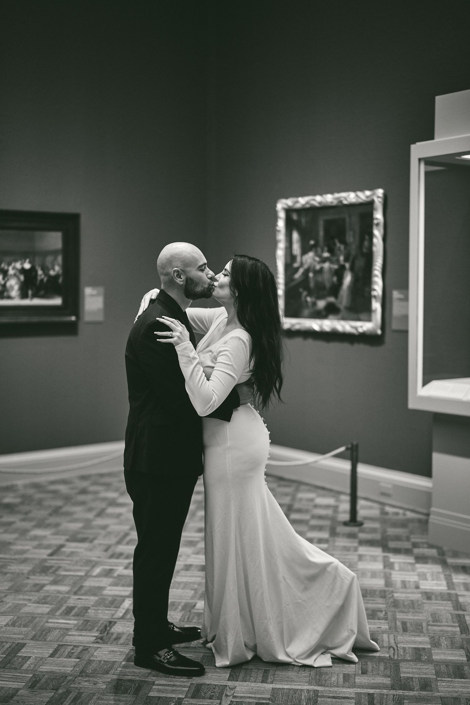 A romantic black and white photo of a couple in formal attire kissing in an art gallery with framed paintings on the walls.