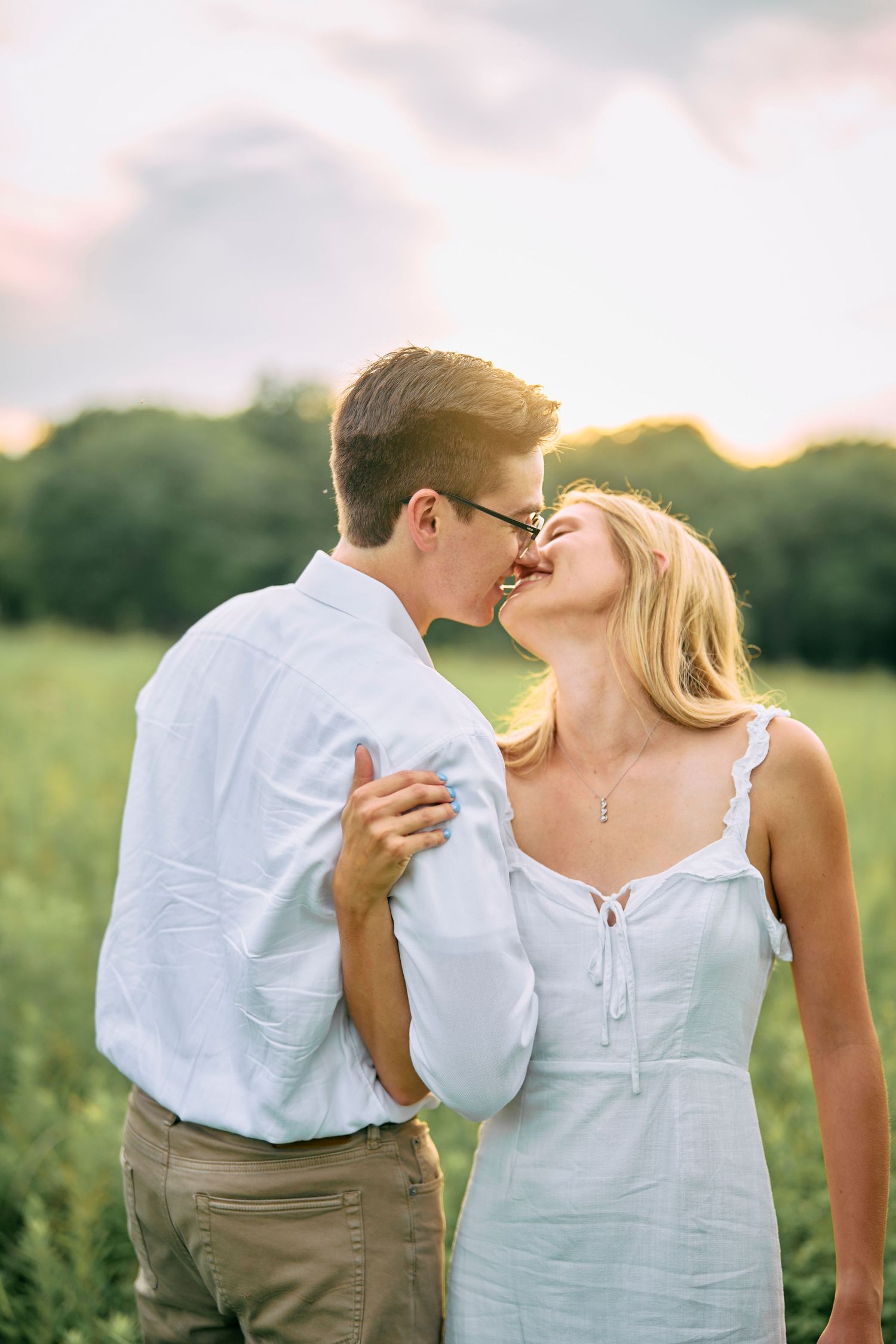 A romantic couple in white outfits share an intimate moment while dancing outdoors at sunset in a grassy field.