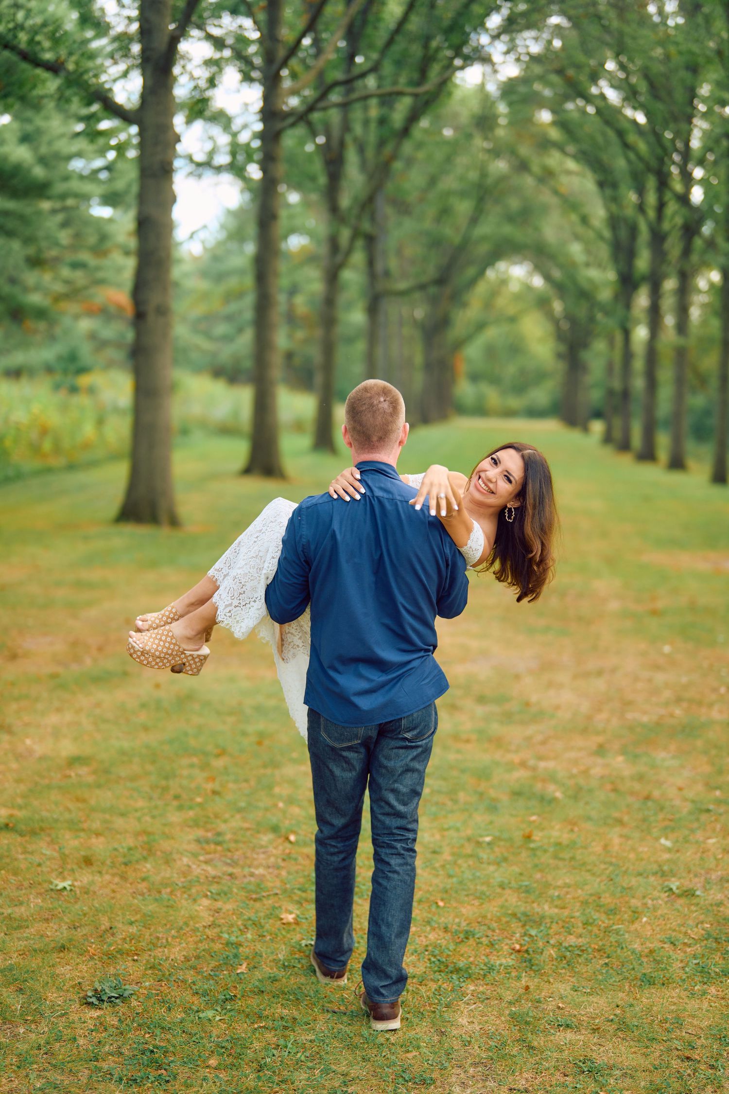 A couple shares a romantic moment on a tree-lined path as one carries the other through a lush green park.