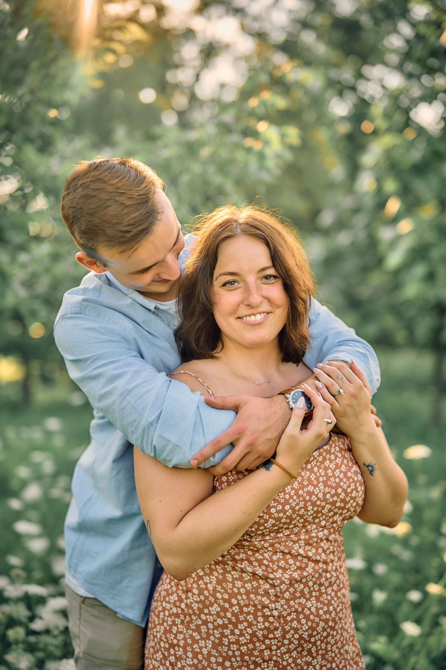 A romantic couple embraces in a sun-dappled garden setting wearing casual summer attire against lush green foliage.