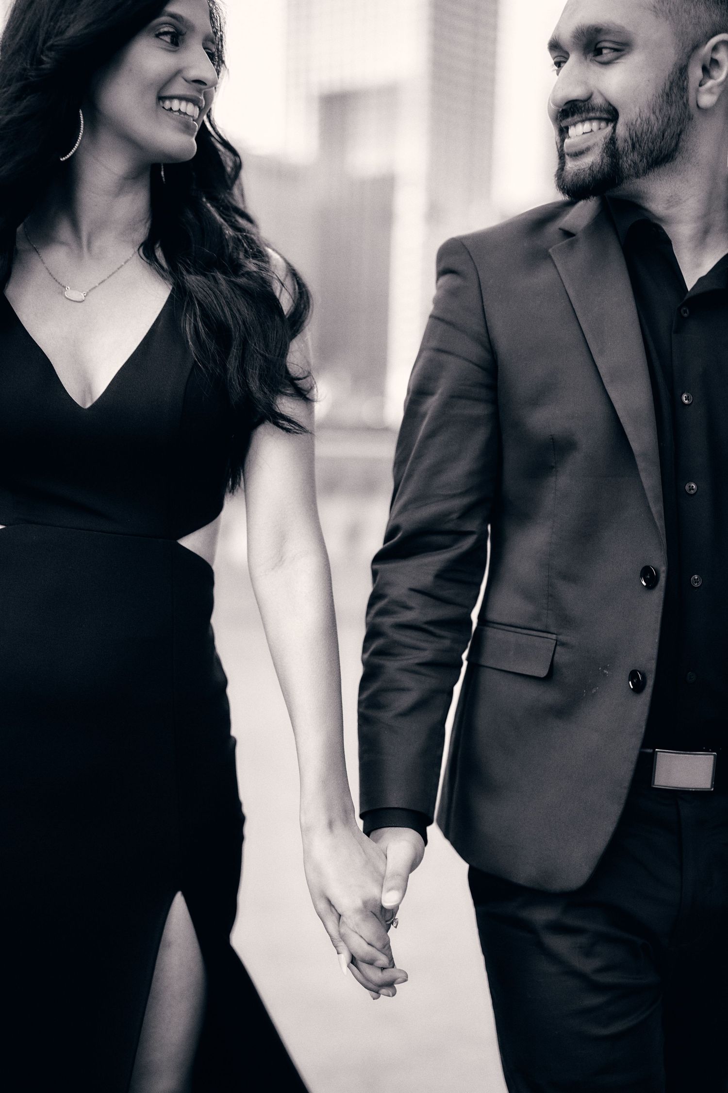 Black and white portrait of couple in formal wear holding hands with shared smiles and urban backdrop.