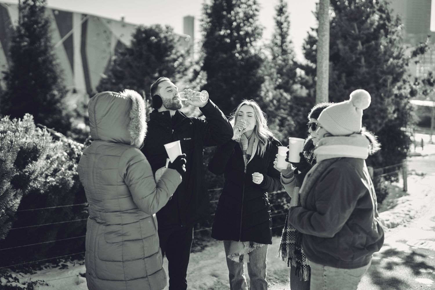 A black and white photo of people in winter coats celebrating with drinks outside among evergreen trees.