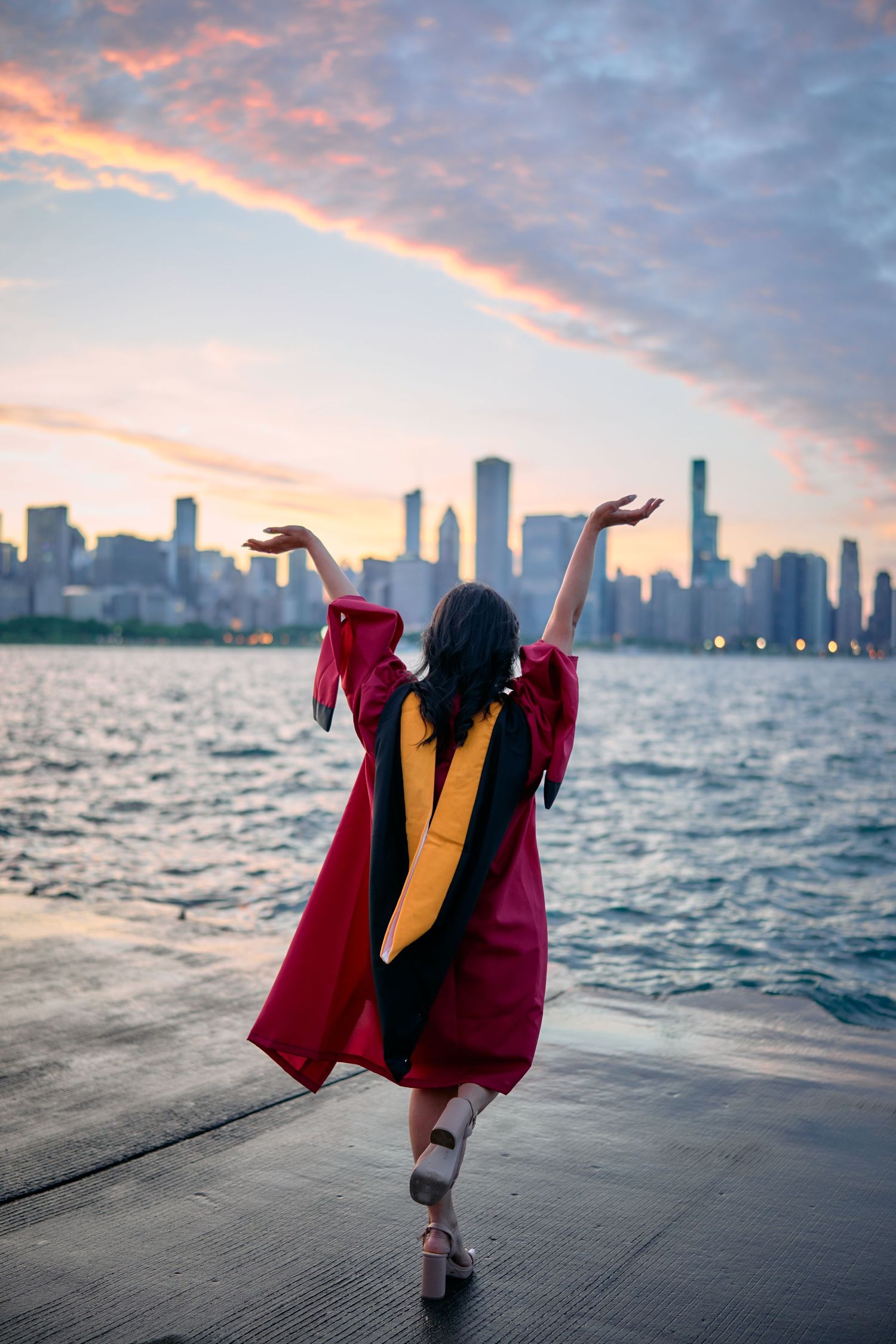 Graduate in red and gold academic regalia celebrates against Chicago skyline at sunset over Lake Michigan.
