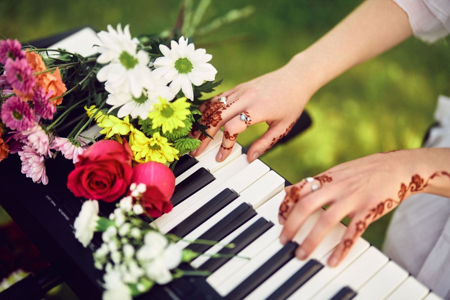Hands adorned with henna play piano keys decorated with colorful daisies, roses and mixed flowers in an outdoor setting.