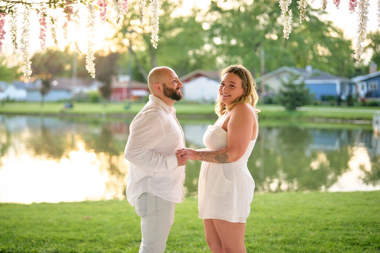 A couple in white outfits stands together on a grassy lawn by a lake at sunset, with wisteria blooming overhead.