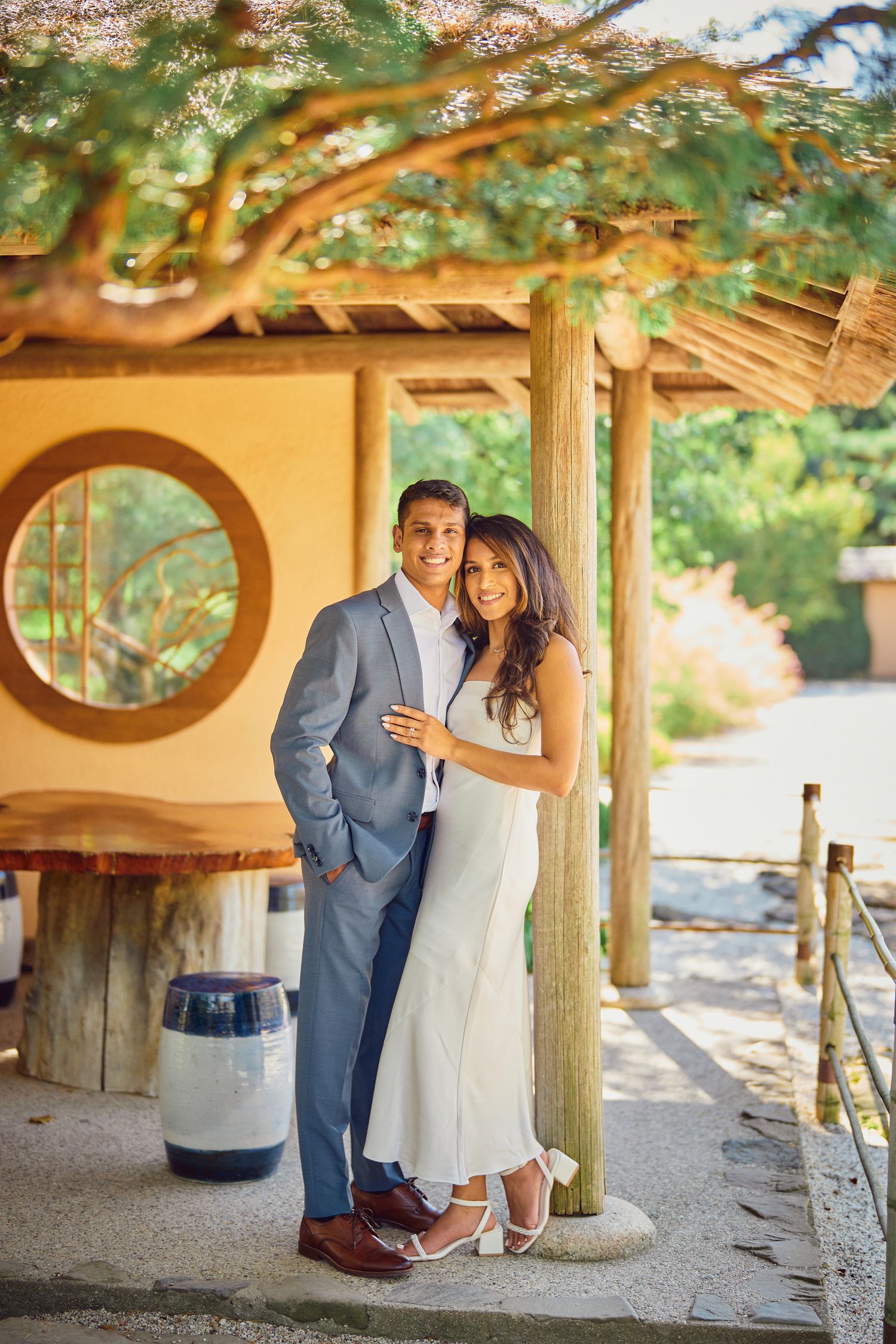 A couple poses together under a wooden gazebo with a circular window and rustic architecture in a garden setting.