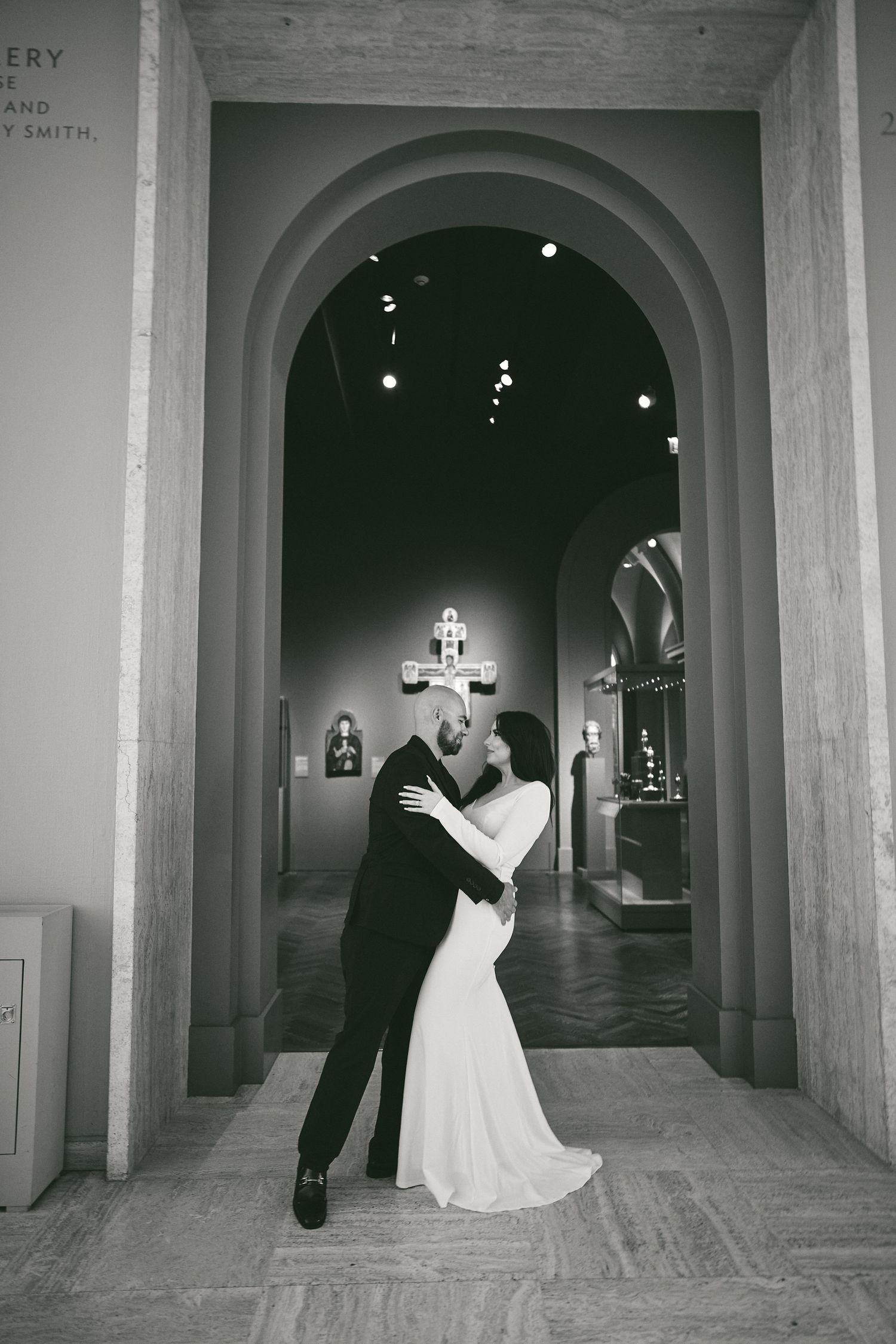 A romantic black and white photo showing a couple embracing in an elegant arched hallway of a historic building.