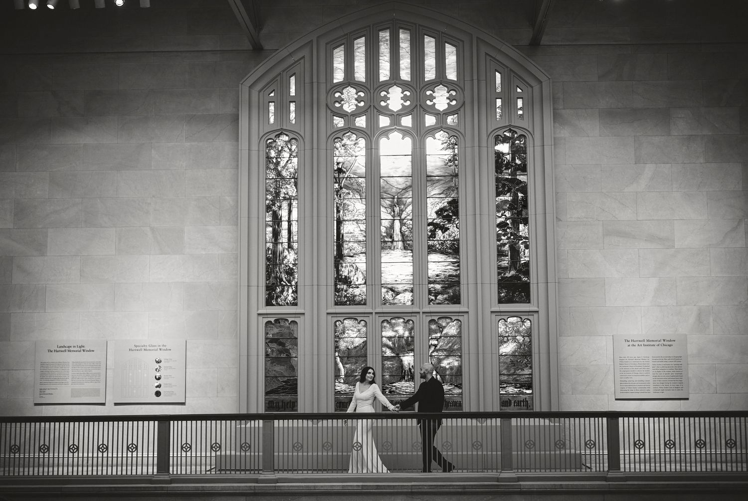 Two people sit in silhouette on a railing in front of a large ornate stained glass window in a church or chapel.